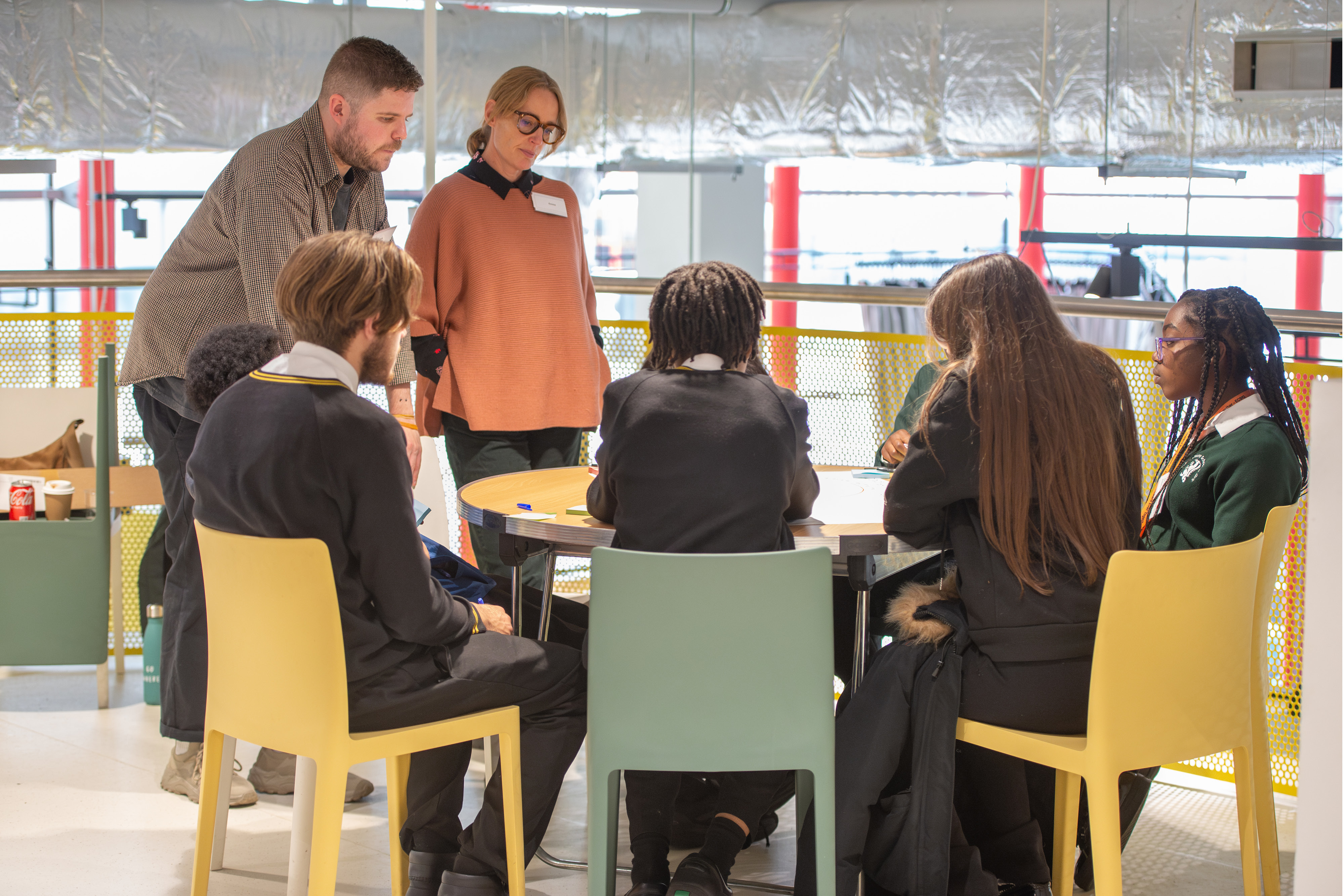 Two adults stand with a group of students seated around a table in a bright, modern room. They appear to be engaged in a discussion or group activity.