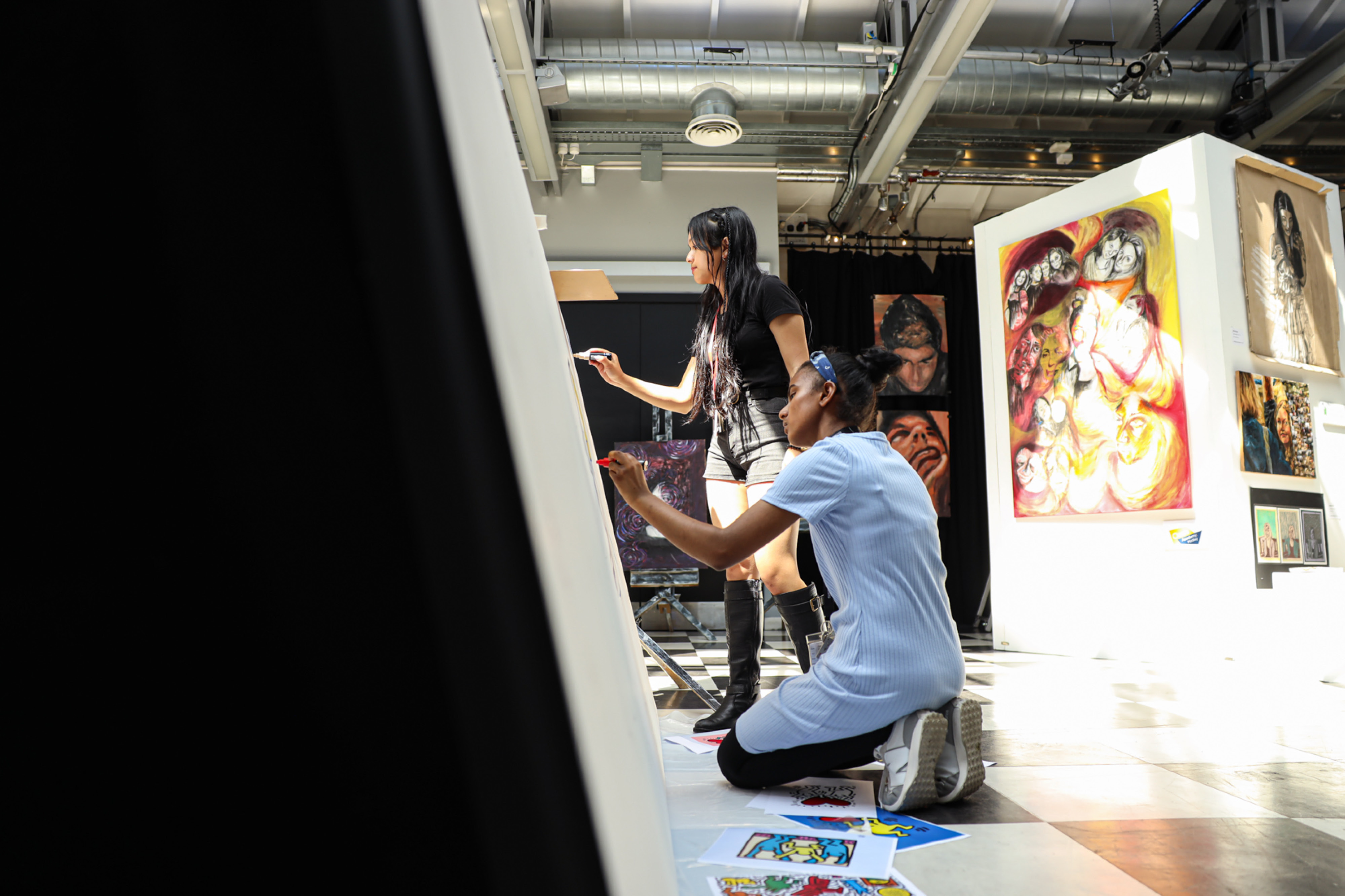 Two people work together on a large art canvas in a well-lit studio or gallery. One stands painting while the other kneels on the floor drawing, surrounded by colorful artworks on the walls and floor.
