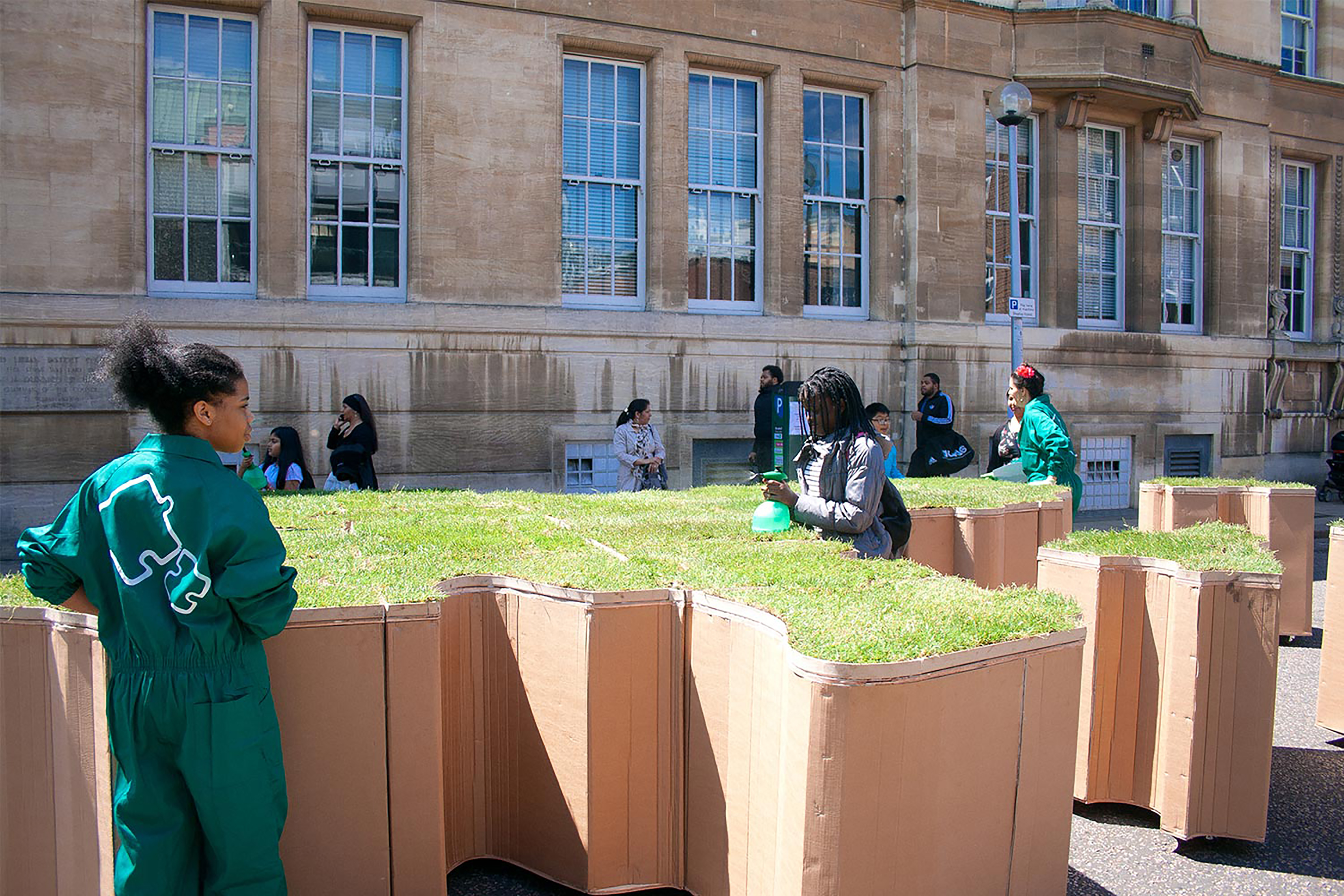 A group of people walking past the large terracotta jigsaw puzzle shaped turf blocks in front of a stone building.