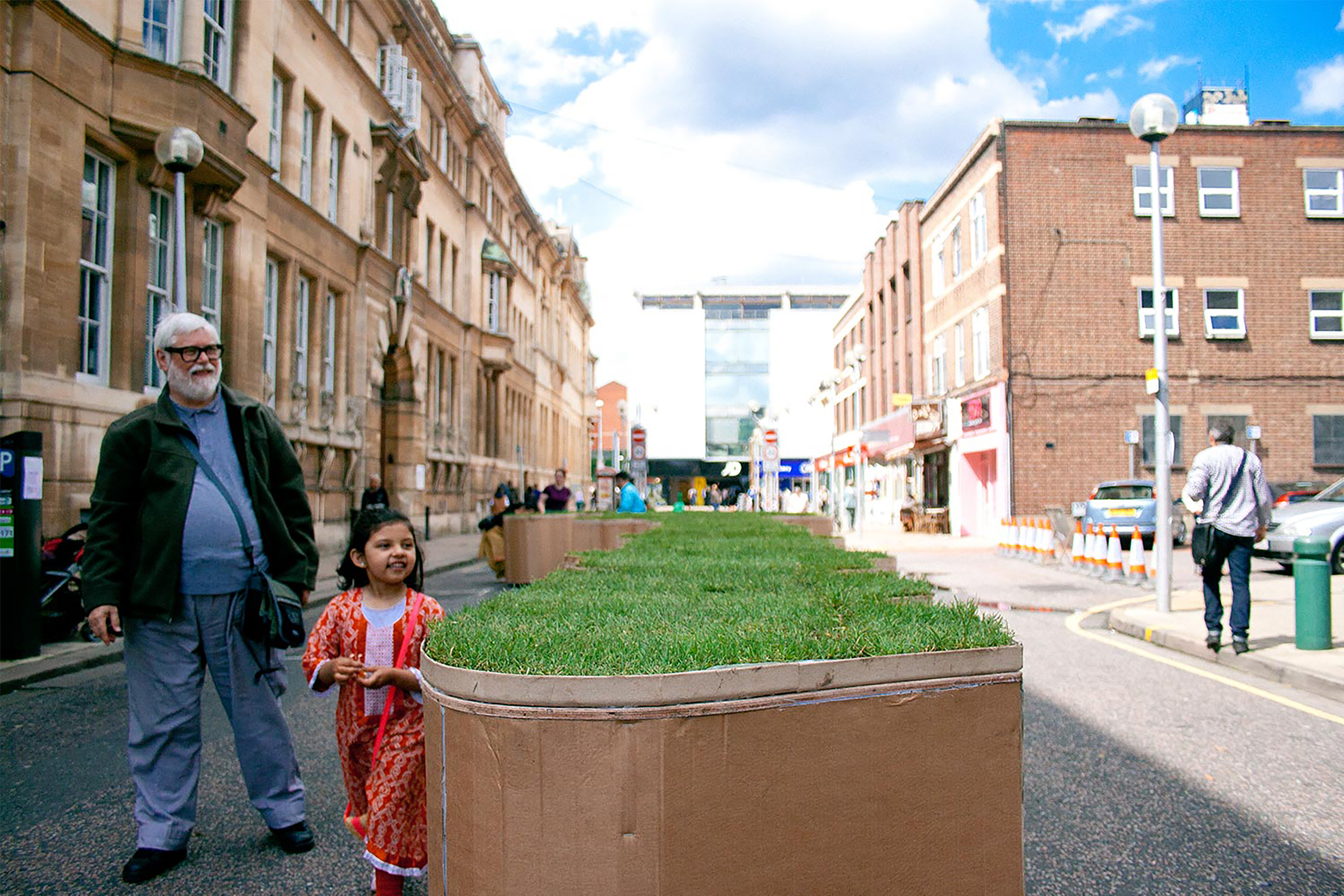 An elderly man and girl smiling standing next to a large terracotta jigsaw puzzle shaped turf block in the middle of a street.