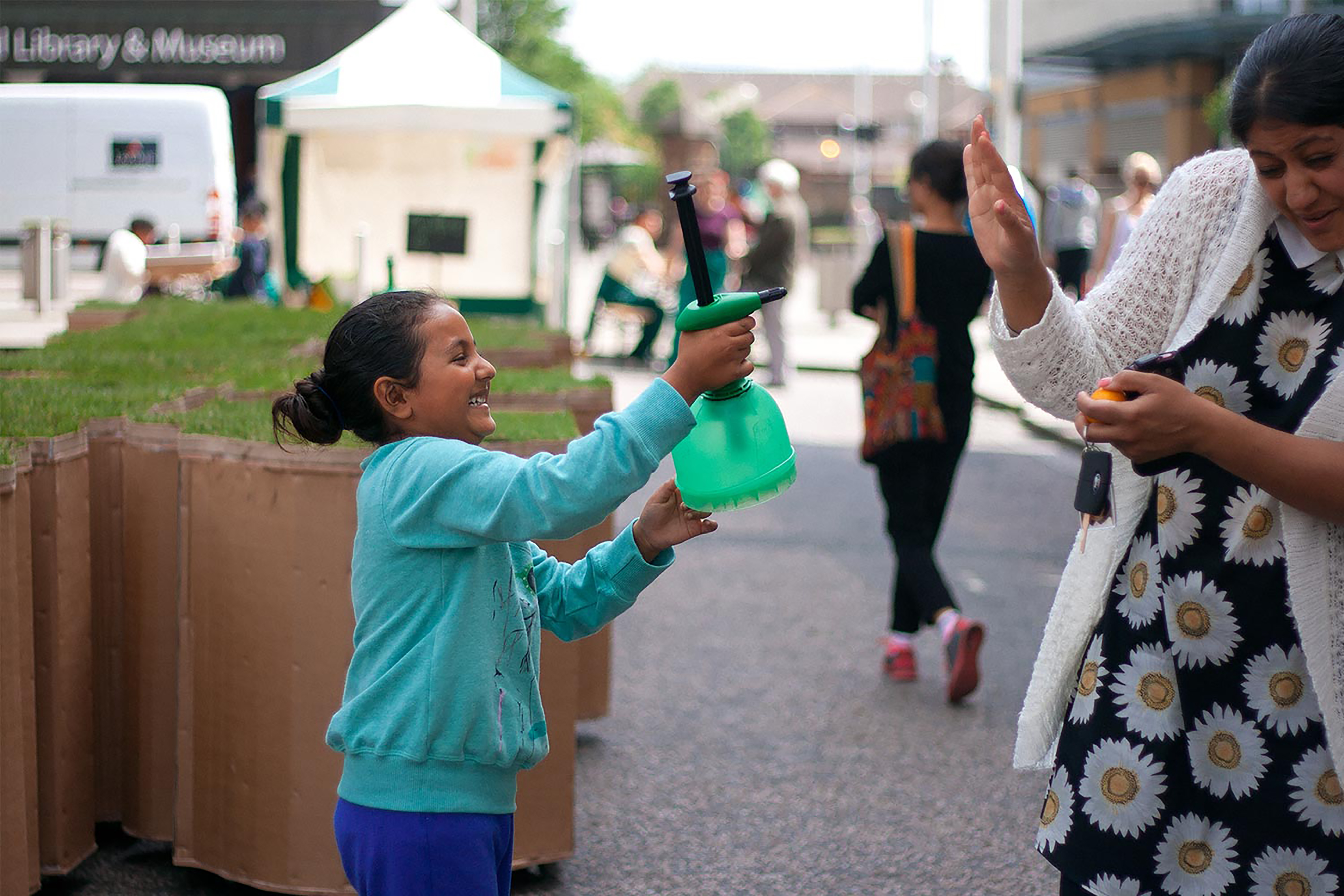 A child playfully spraying her mother with a green garden spray bottle in front of a large terracotta jigsaw puzzle shaped turf block.