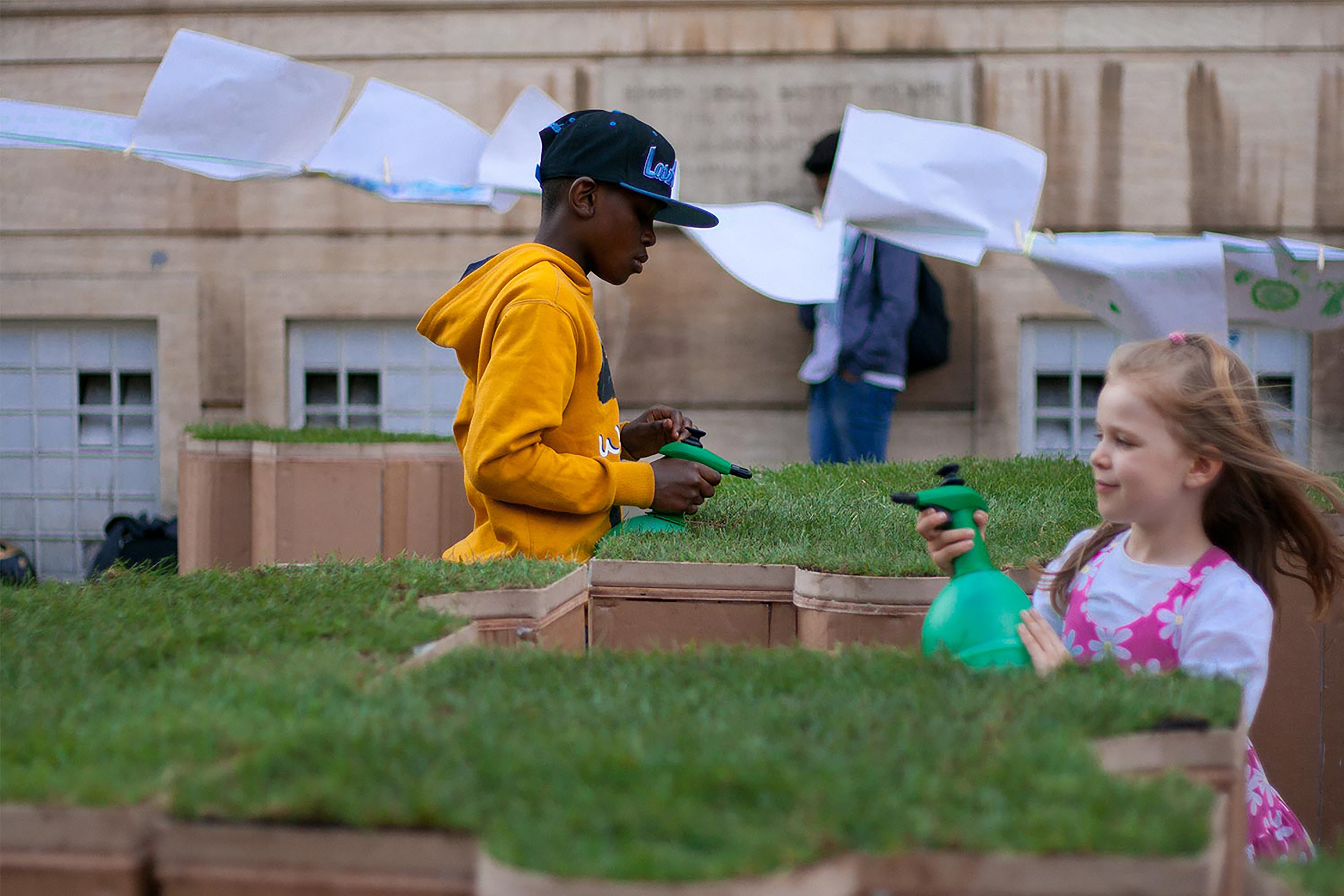 Two children using green garden spray bottles on a patch of turf on top of a large terracotta jigsaw puzzle shaped turf block.