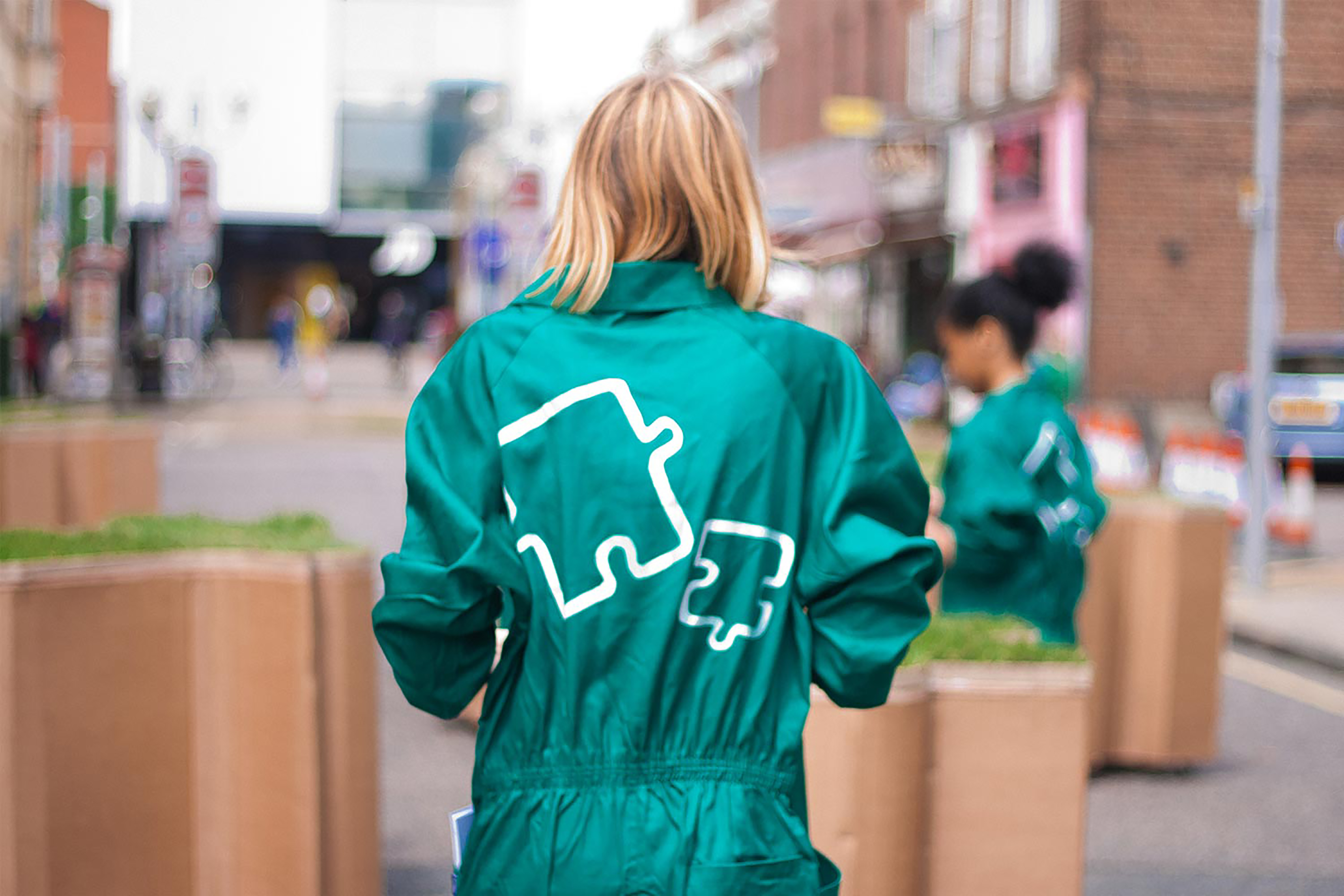 A woman wearing a green jumpsuit with the outline of a jigsaw puzzle piece on the back walking through large terracotta jigsaw puzzle shaped turf blocks.
