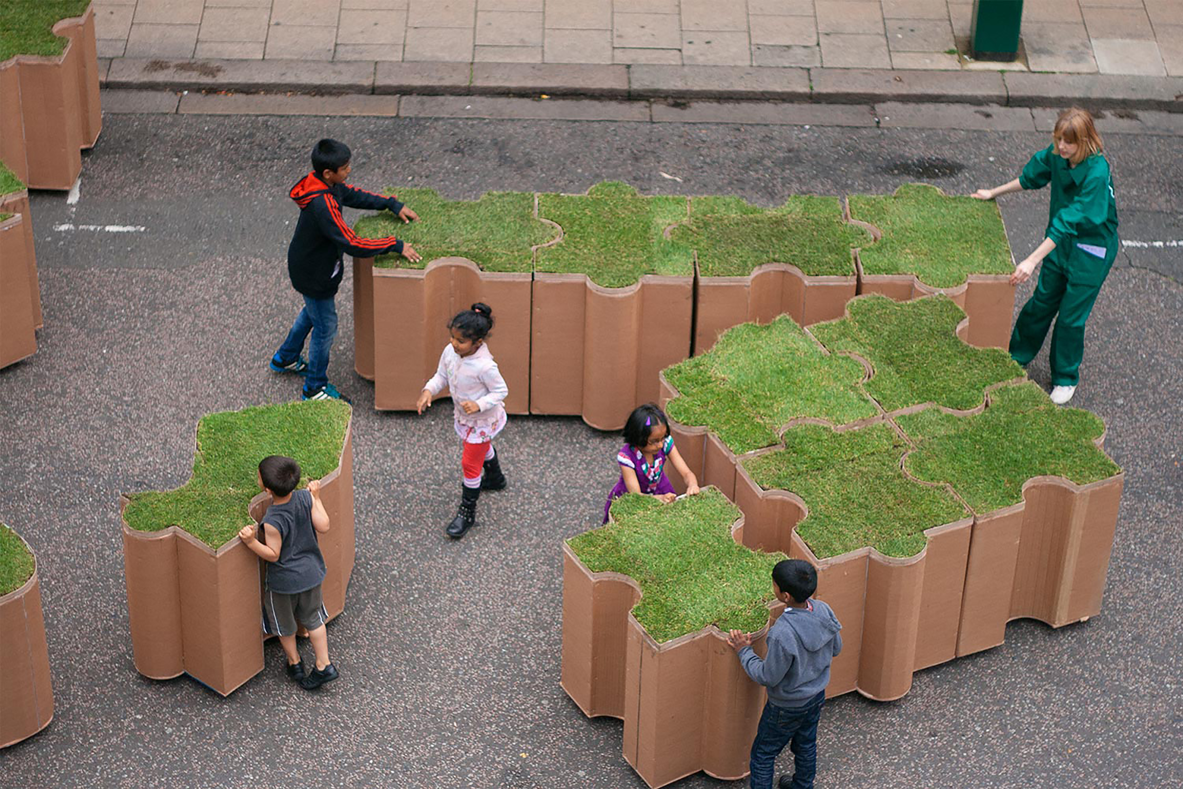 A group of children and a gardener assembling several large terracotta jigsaw puzzle shaped turf blocks.