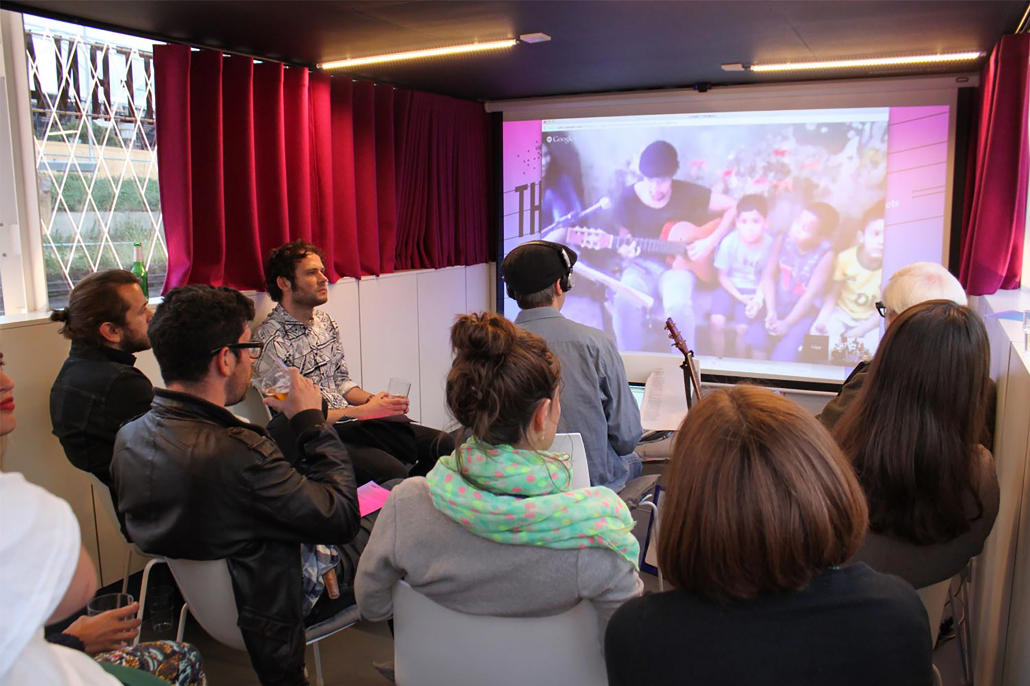 A group of people in a small room with purple curtains all watching a small projection screen showing a film.