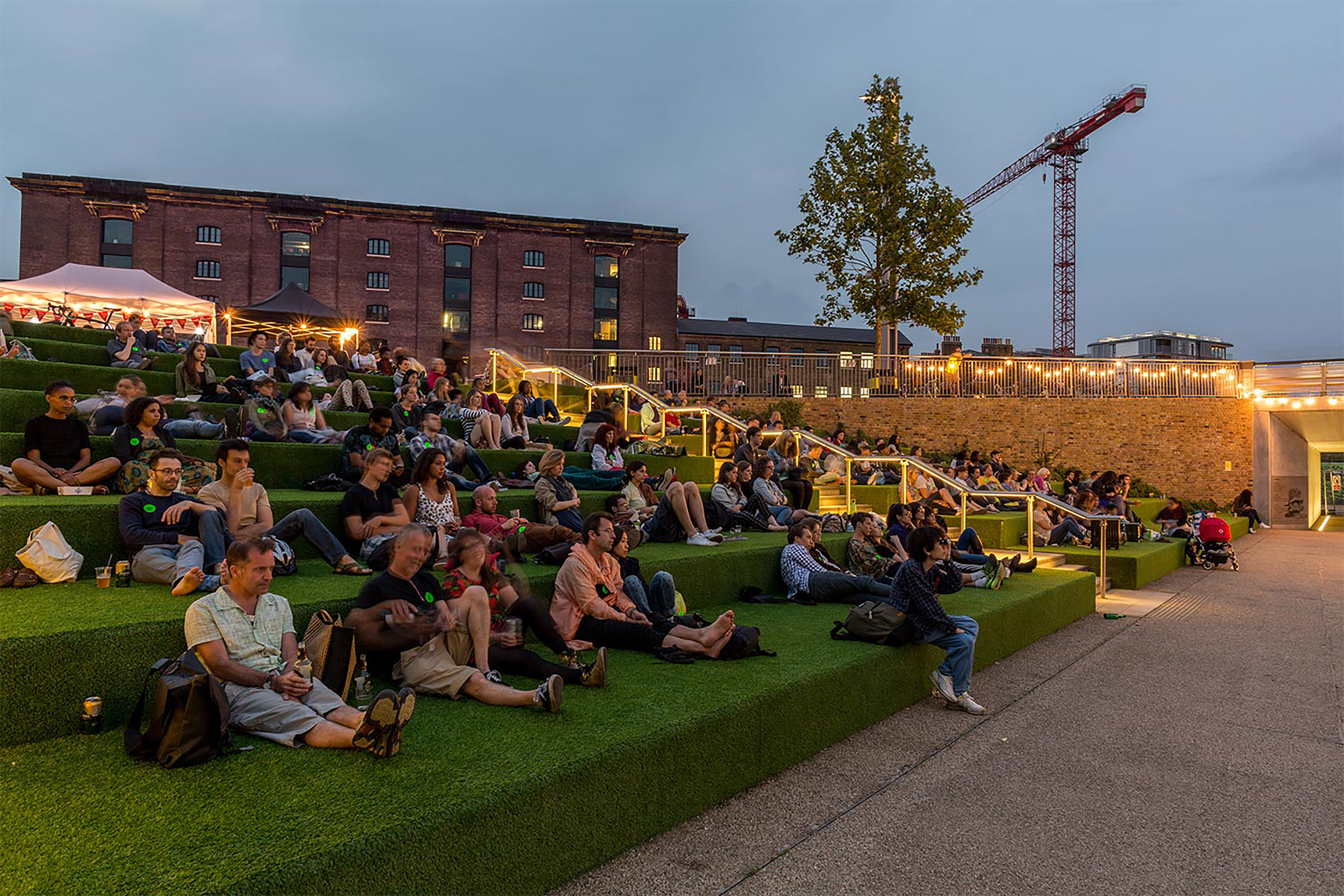 A large group of people at dusk all sitting on the ground facing towards a projection screen.