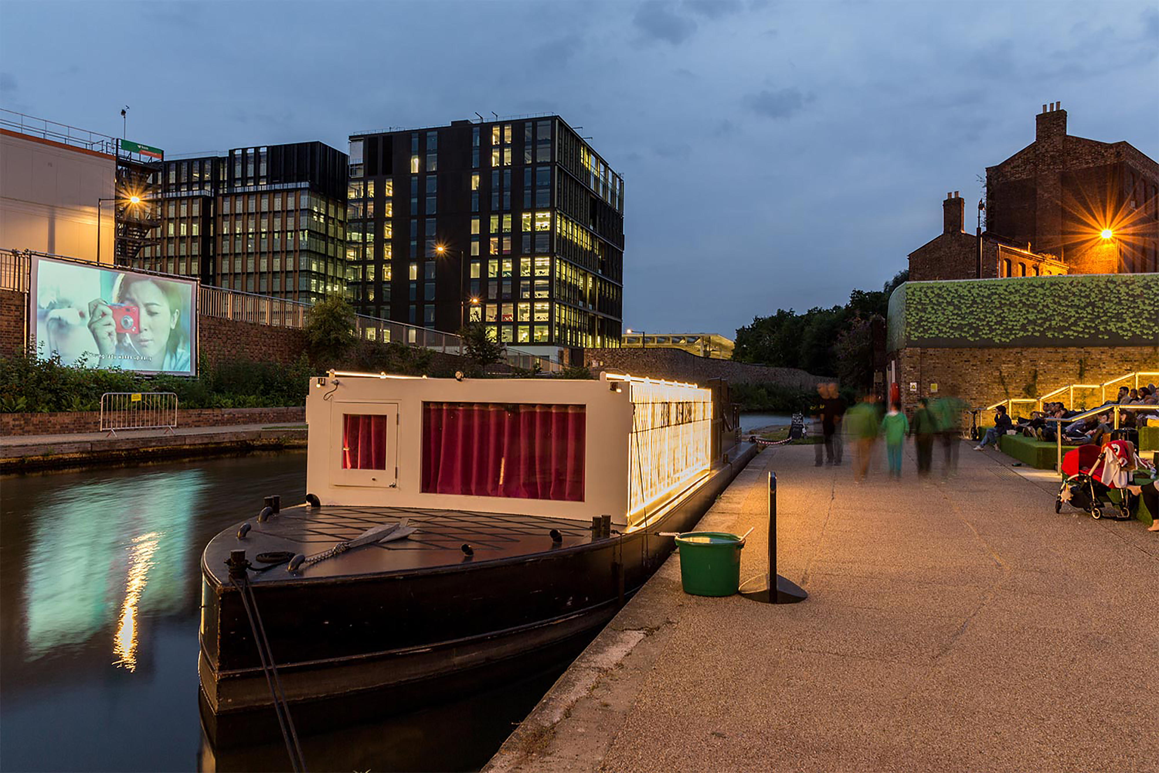 A large canal boat parked up next to a bank with people watching a large projection screen on the other side of the water.
