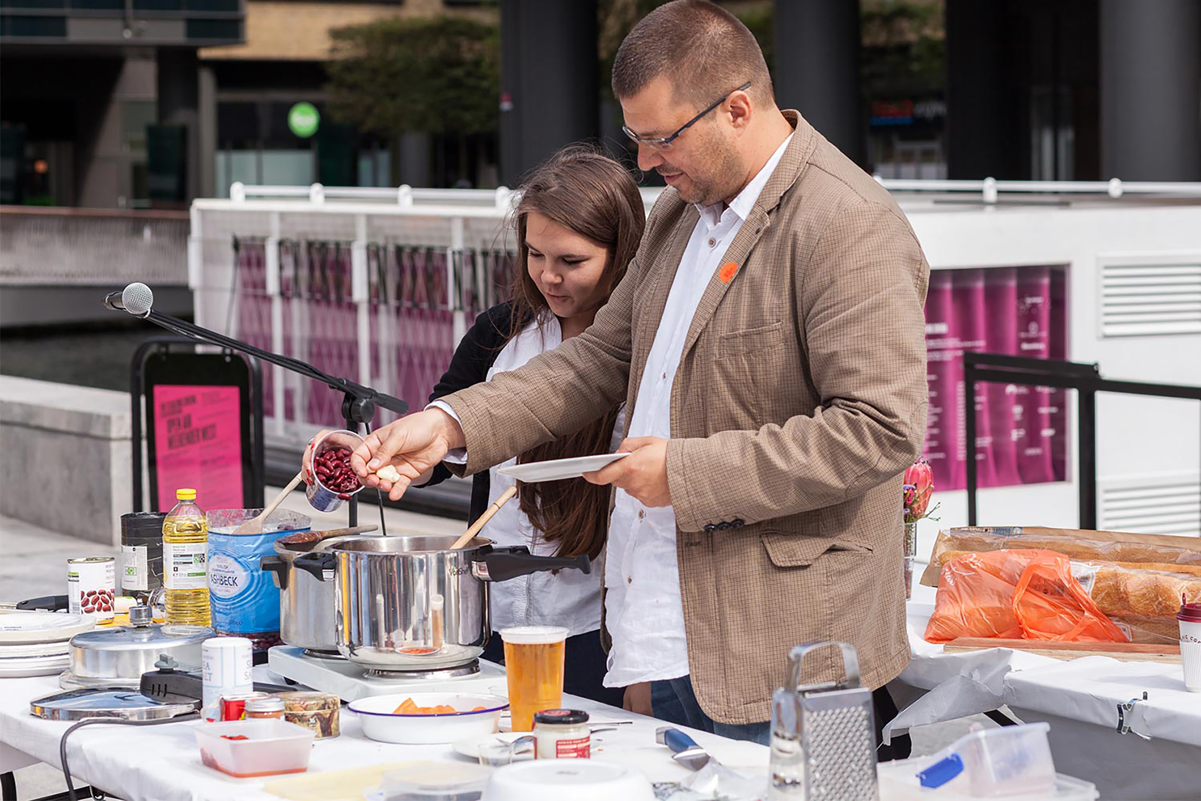 Two people standing behind a large white table full of different food and drink products, with a large boat in the background.
