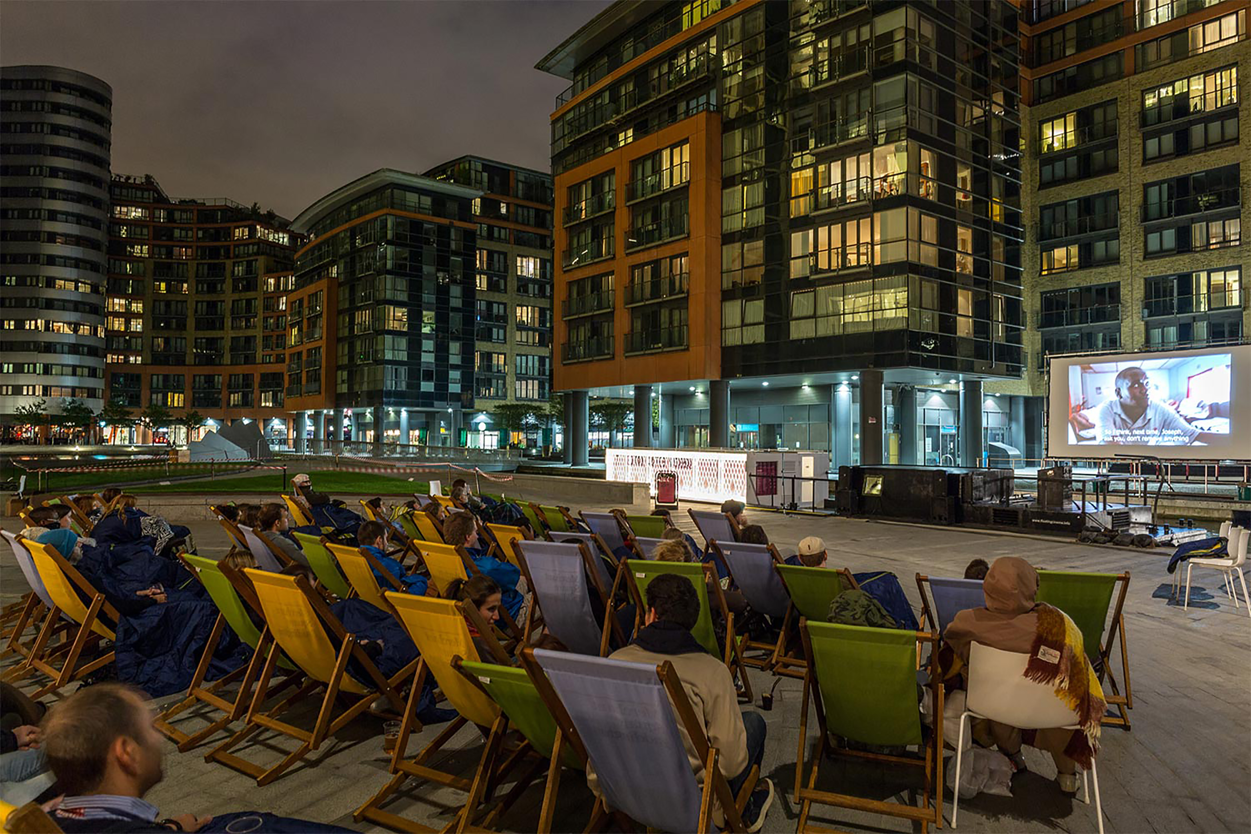 A large group of people at night all sitting on chairs facing towards a projection screen.