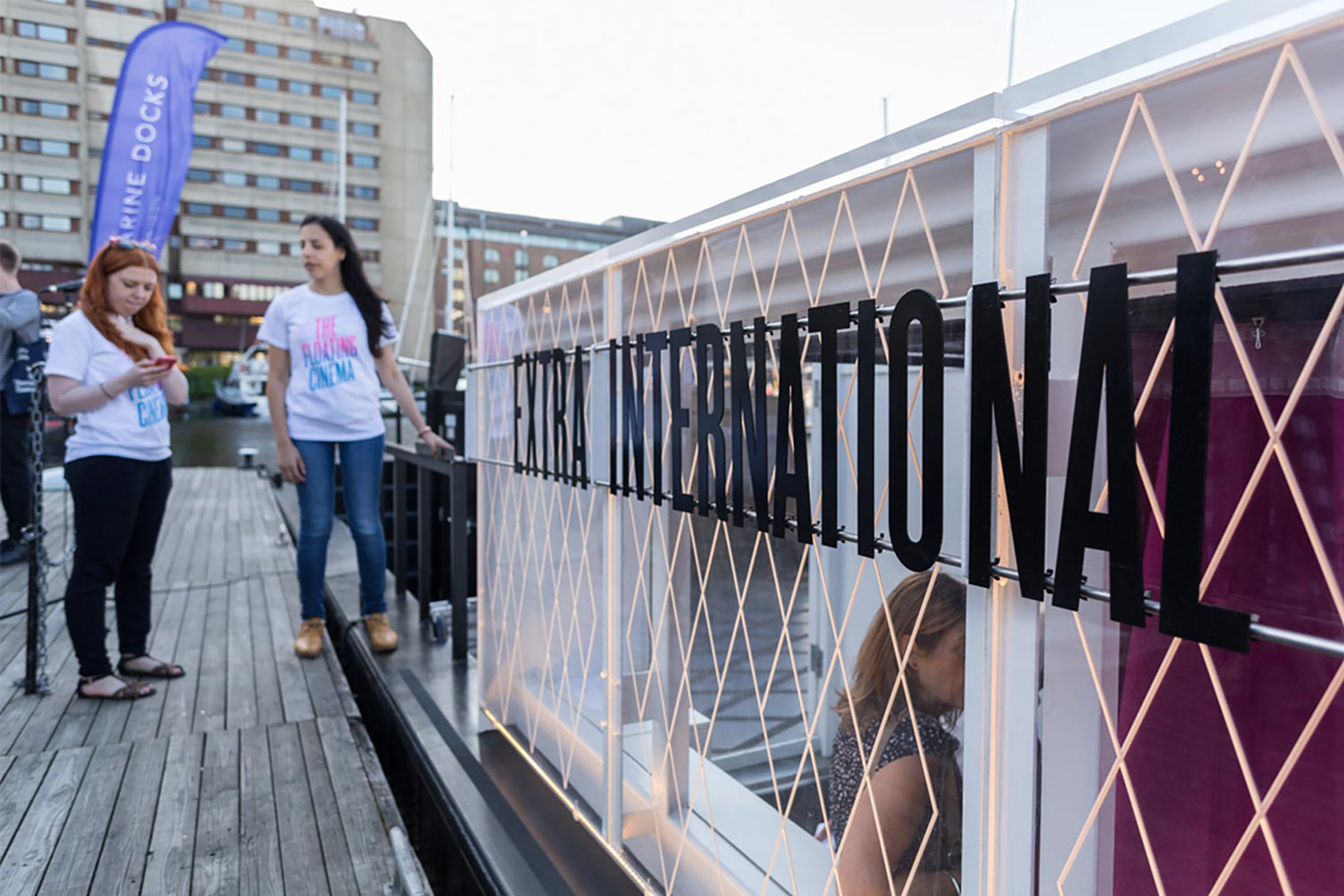 Two women standing on a bank next to a large canal boat with a sign on the side that reads “Extra International”.