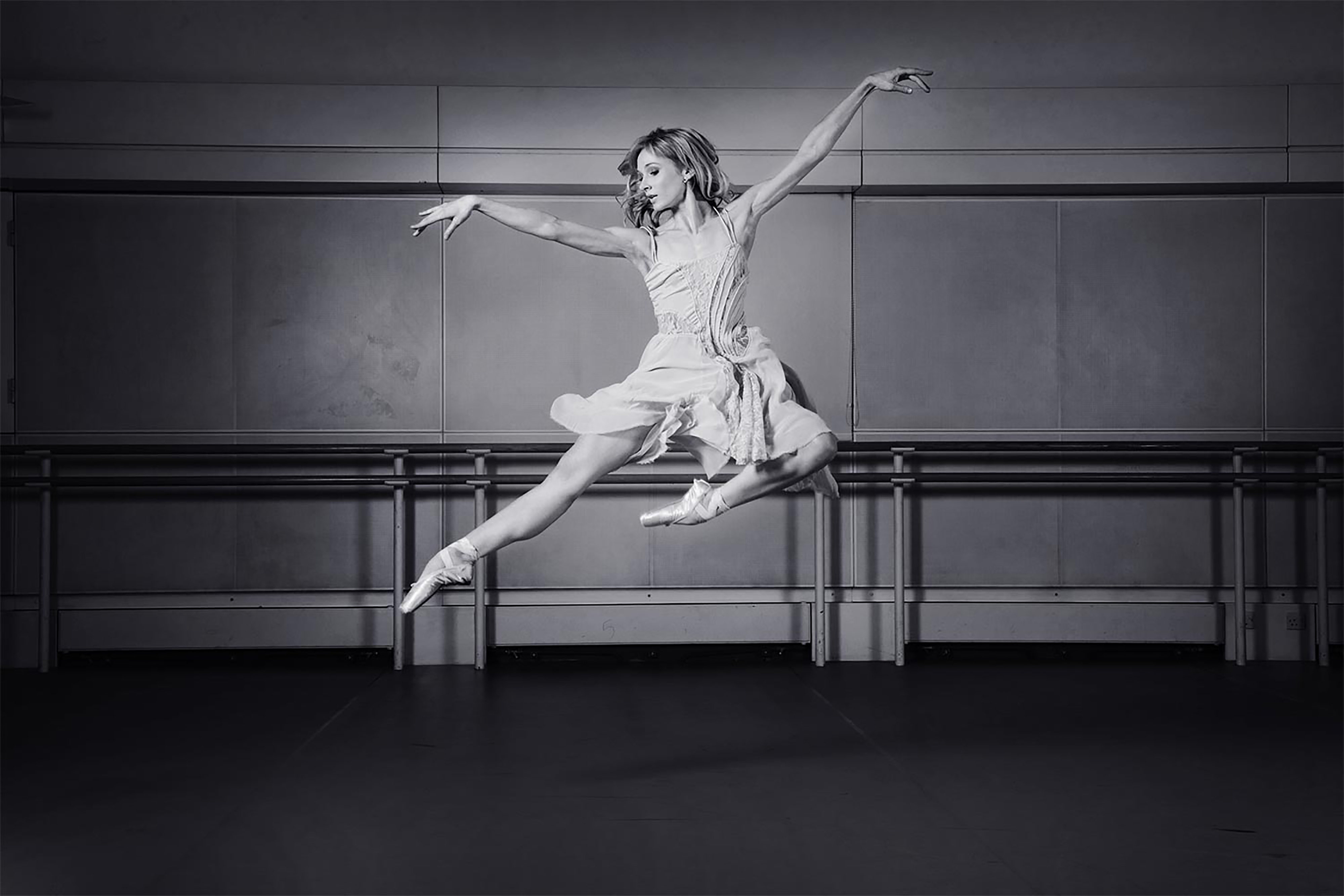 A black and white photograph of a ballerina jumping in the air with her arms out wide, wearing a short white dress, in a dark in front of a balancing beam.