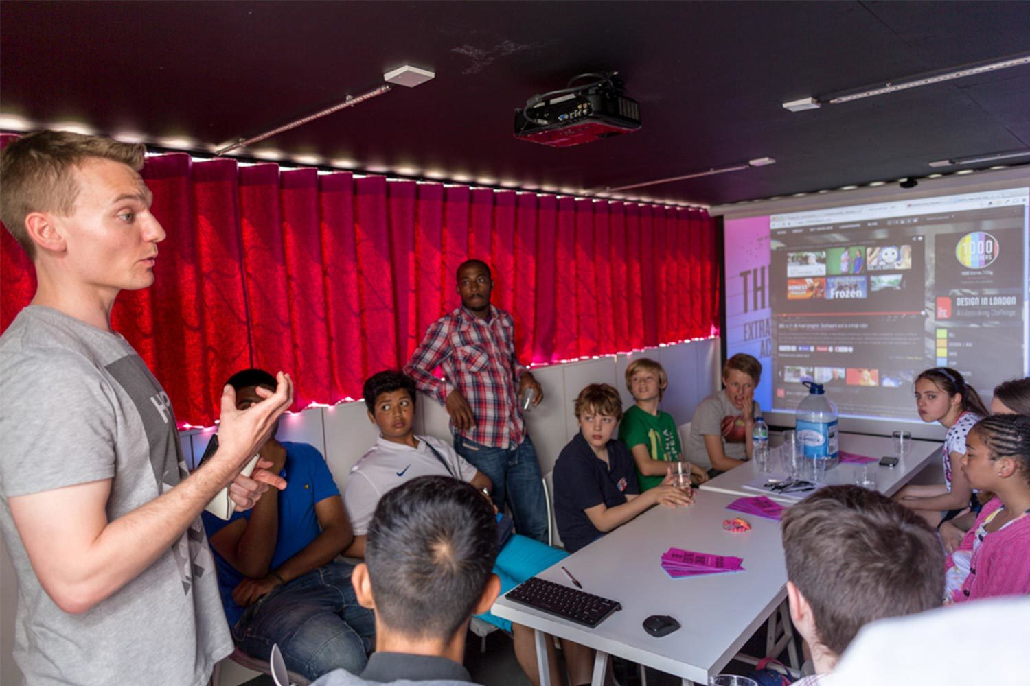 A group of children sitting and talking around a white table with water bottles and pink flyers, in a room with dark red curtains and a man speaking.