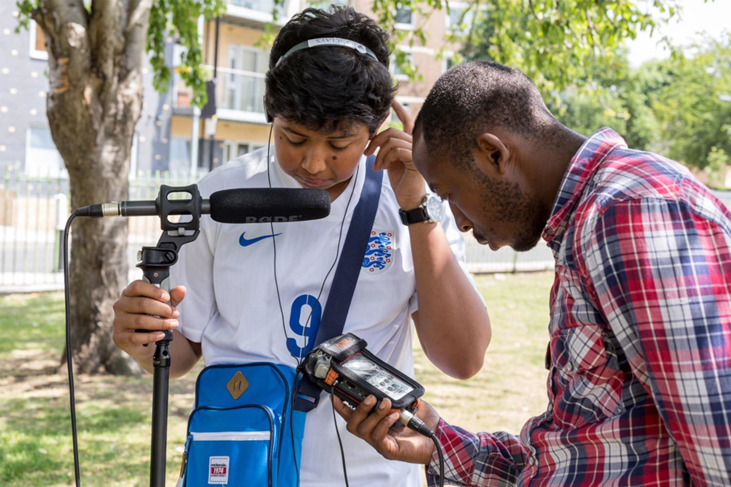 A young boy wearing audio recording equipment speaking into a large microphone next to a man checking an audio recorder.
