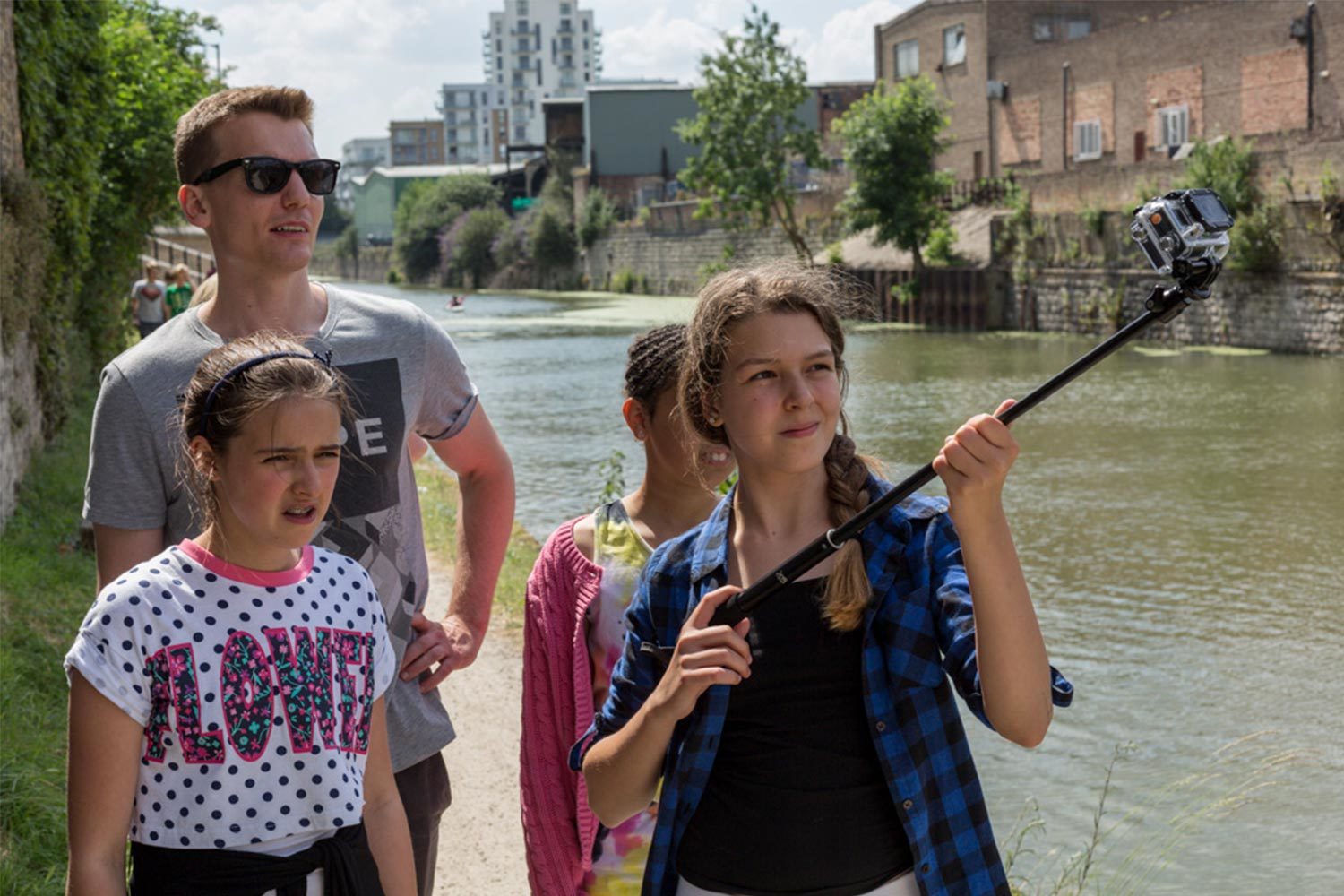 A group of children and adults recording and filming outdoors next to a bank of water.