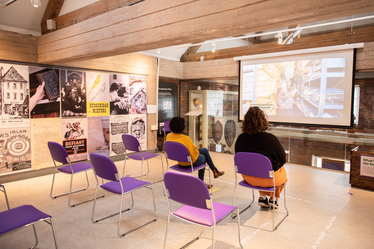 A dim-lit room with rows of purple chairs with two people watching a film screening on a large projector screen.