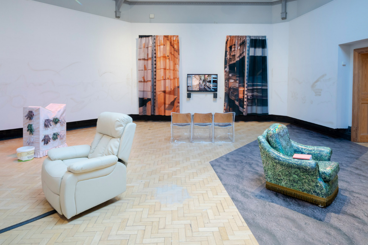 A gallery space containing a row of wooden chair covered by plastic, in front of a tv show a film, with either side displayed with two large printed curtains of shelves; a green patterned chair on a grey flooring; and a cream massage chair next to a wooden block.