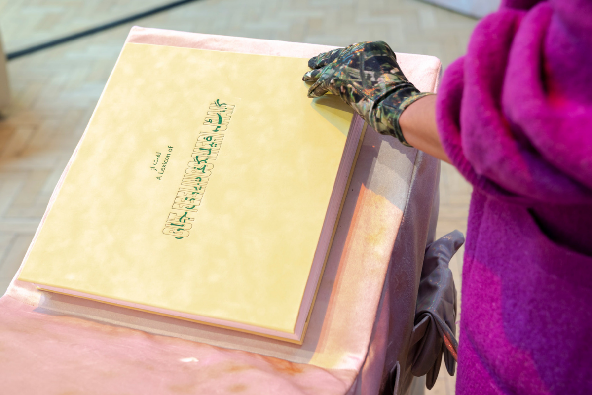 A large yellow velvet book with green title font on a pink plinth being read by a person in a purple jumper and wearing a patterned glove.