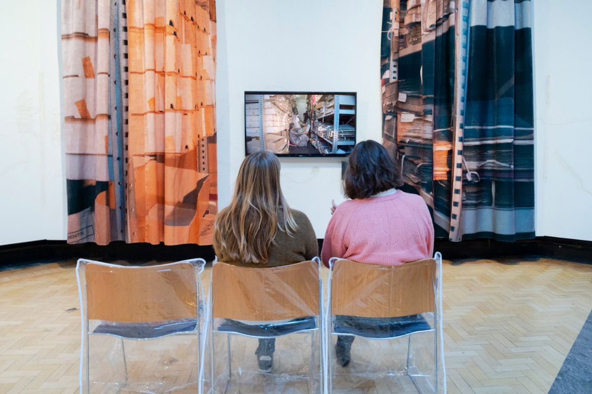 Two people sitting in a row of wooden chair covered by plastic, in front of a tv show a film, with either side displayed with two large printed curtains of shelves.