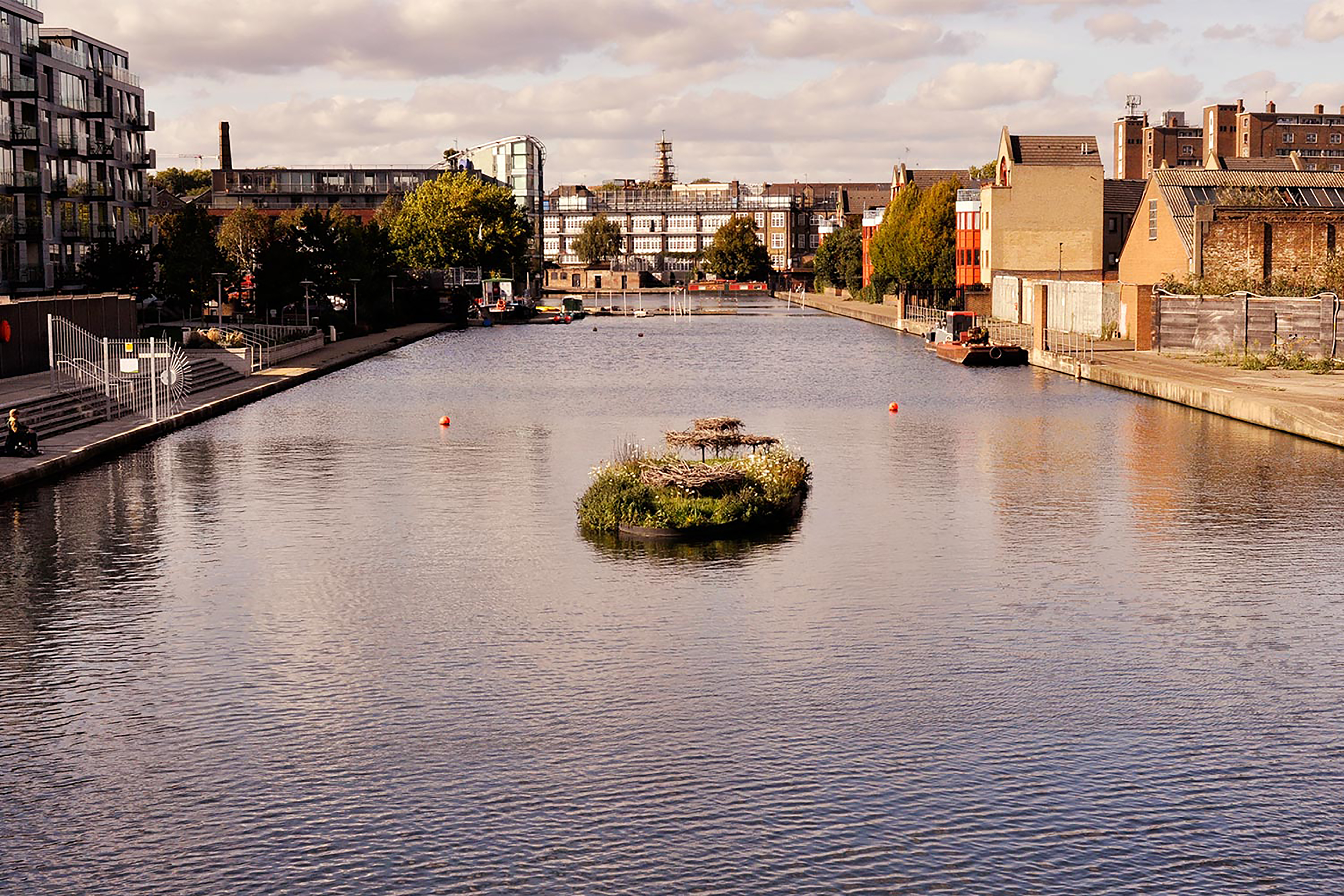 A small patch of grass with white flowers and raised circles of twigs floating on a small canal of water surrounding by stone steps and dark brick buildings.