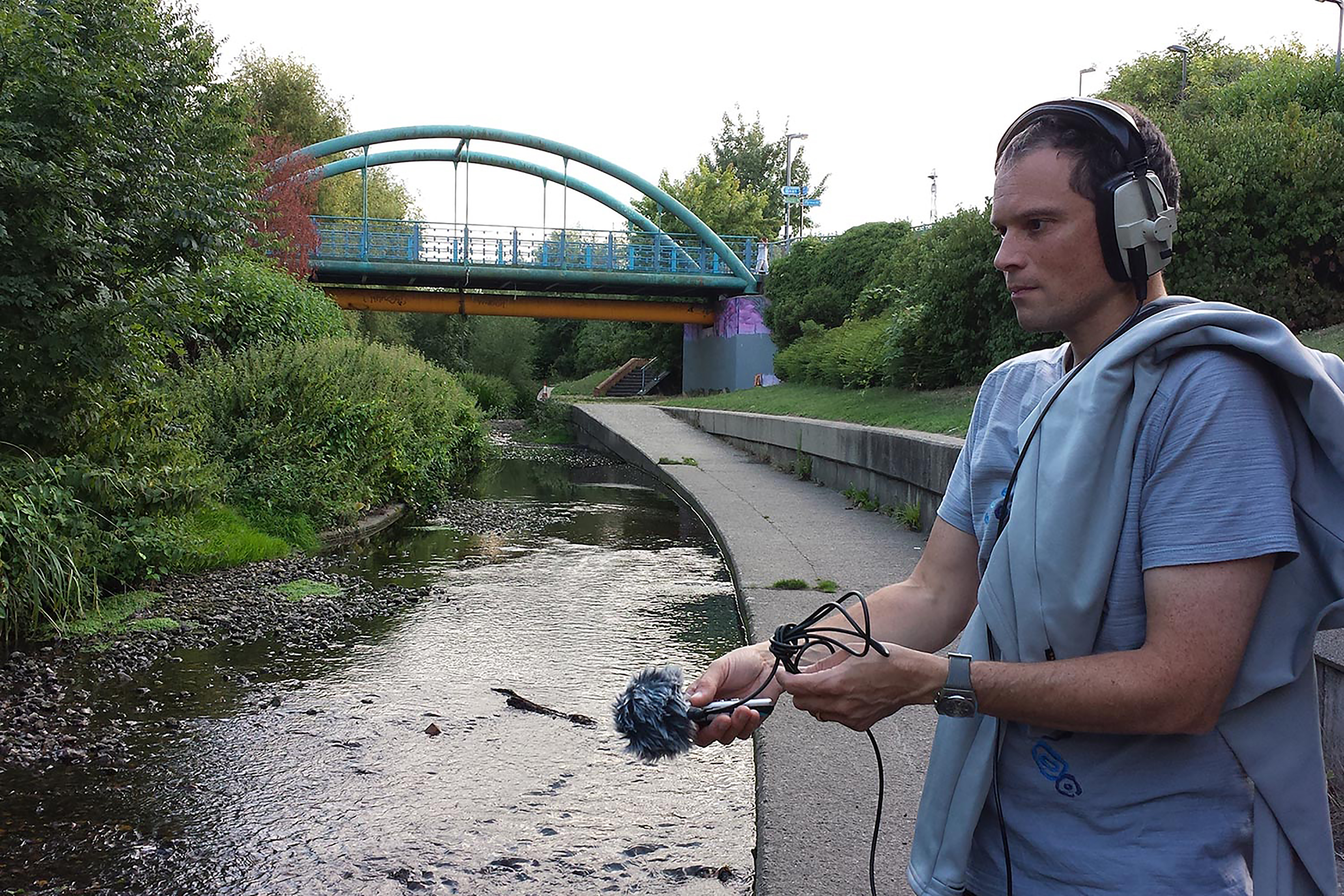 A photograph of a white man wearing headphones and a blue top, operating a microphone beside a canal with a bridge in the background.