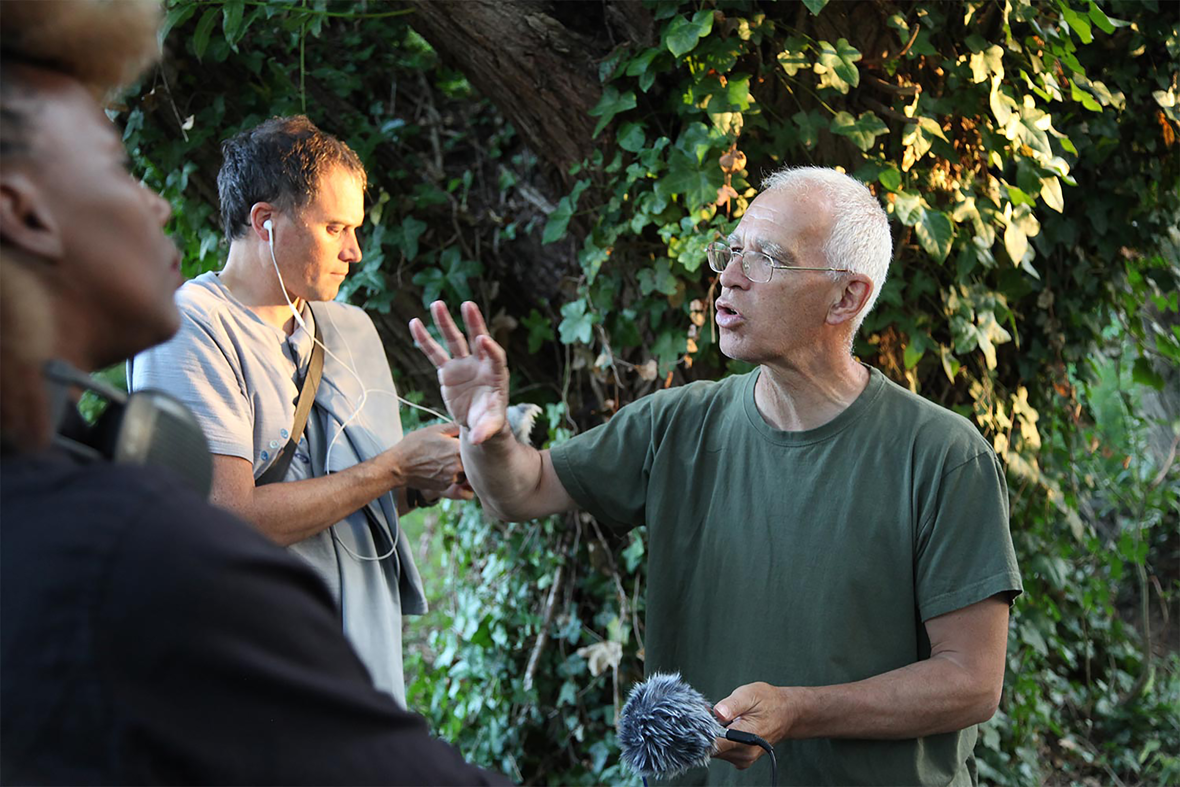 A middle-aged white man wearing a green top and holding a mic while addressing a crowd in front of greenery.