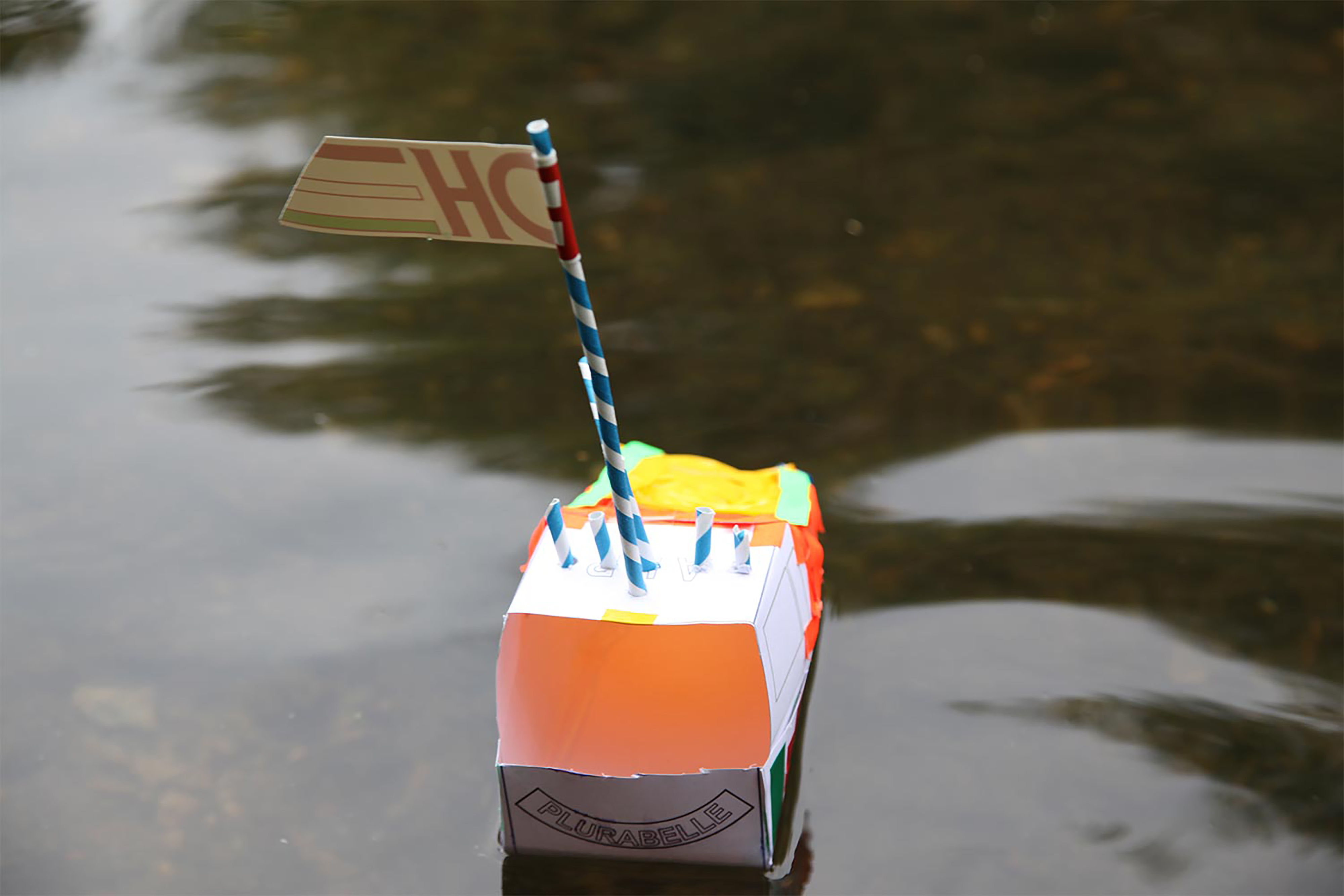 A photograph of a white and orange cardboard boat made with straws floating on the canal.