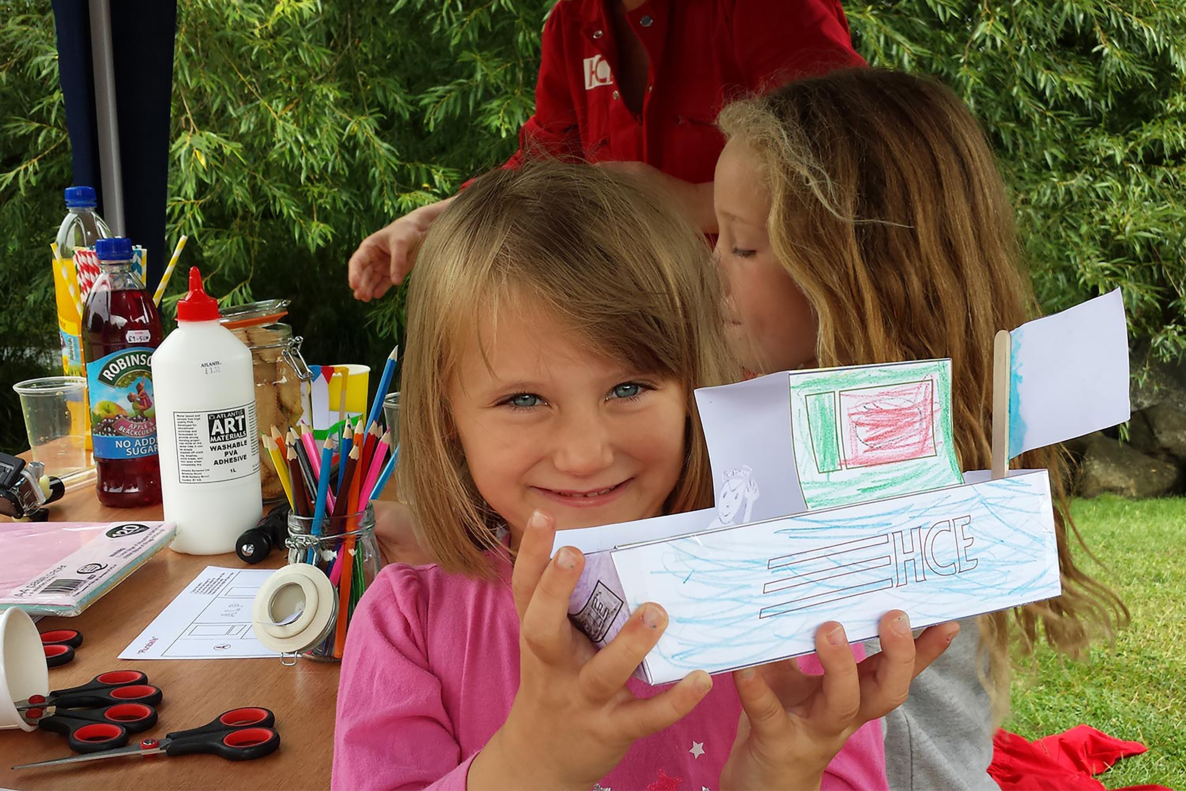 A photograph of a young girl holding a handmade paper boat with the letters HCE marked across.