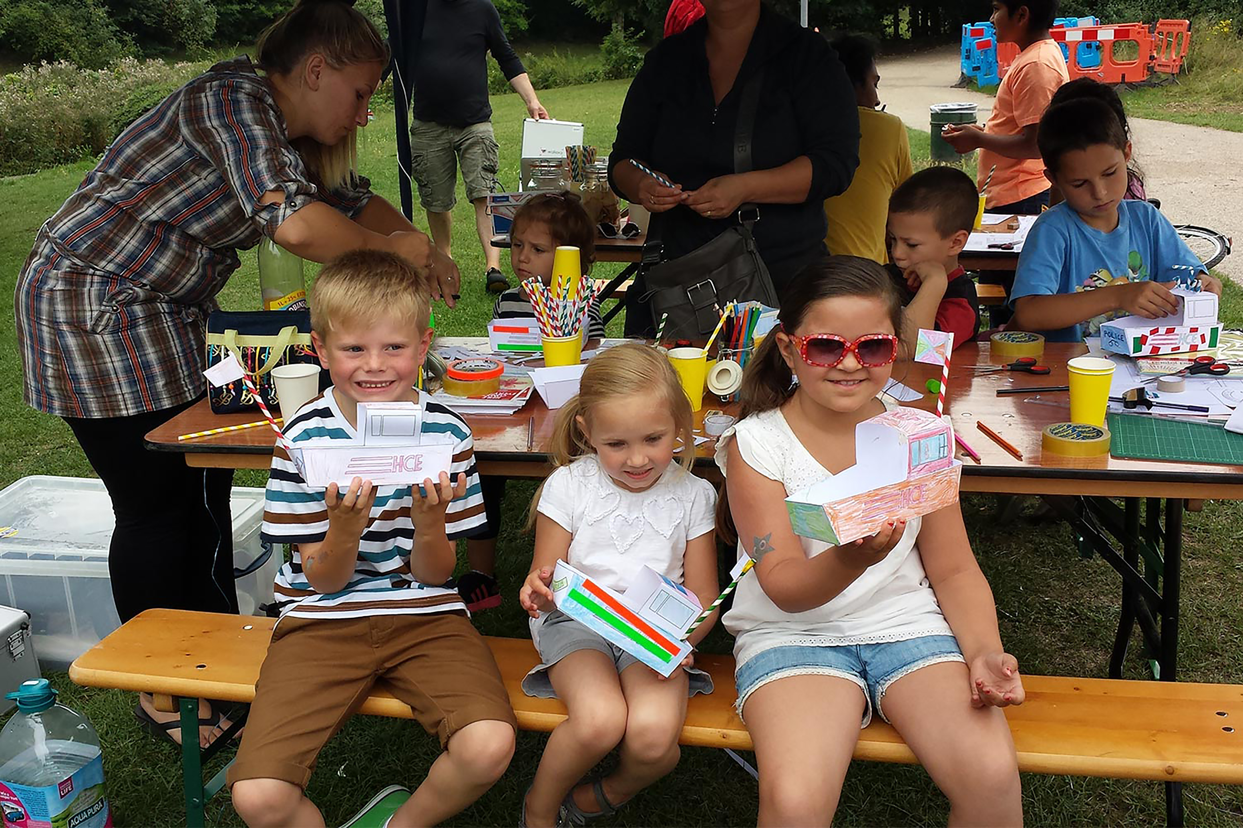 A boy and two girls hold their paper boats sitting on a picnic bench.