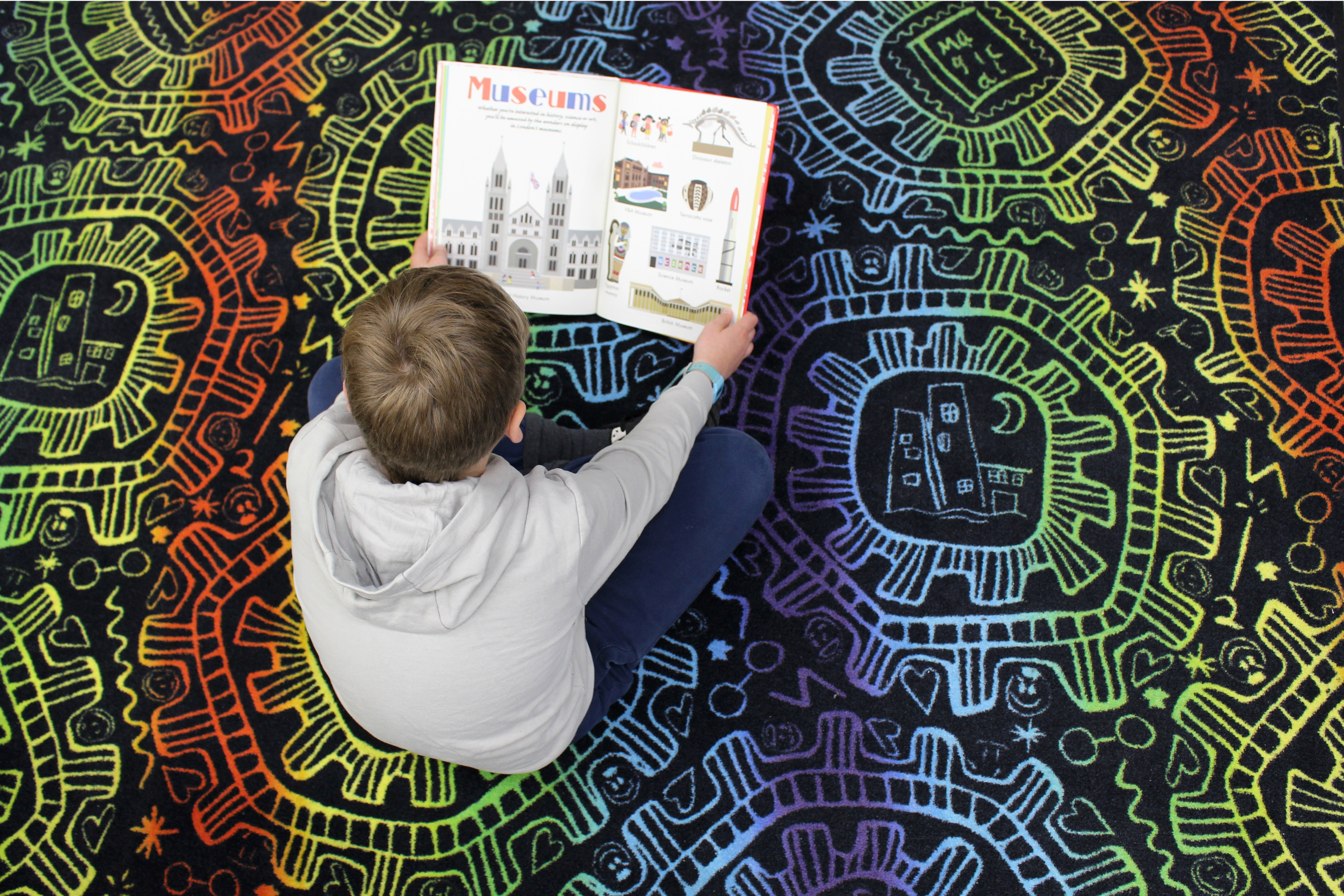 A child sitting on a colourful rainbow patterned rug from above. He is holding a children’s book reading.
