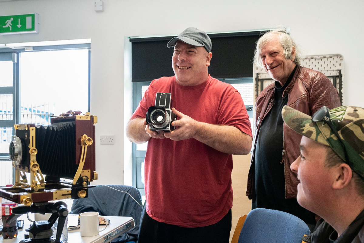 Three men all smiling, with two men looking at another man holding an old black camera, next to a table with a large old camera on in a cream room.