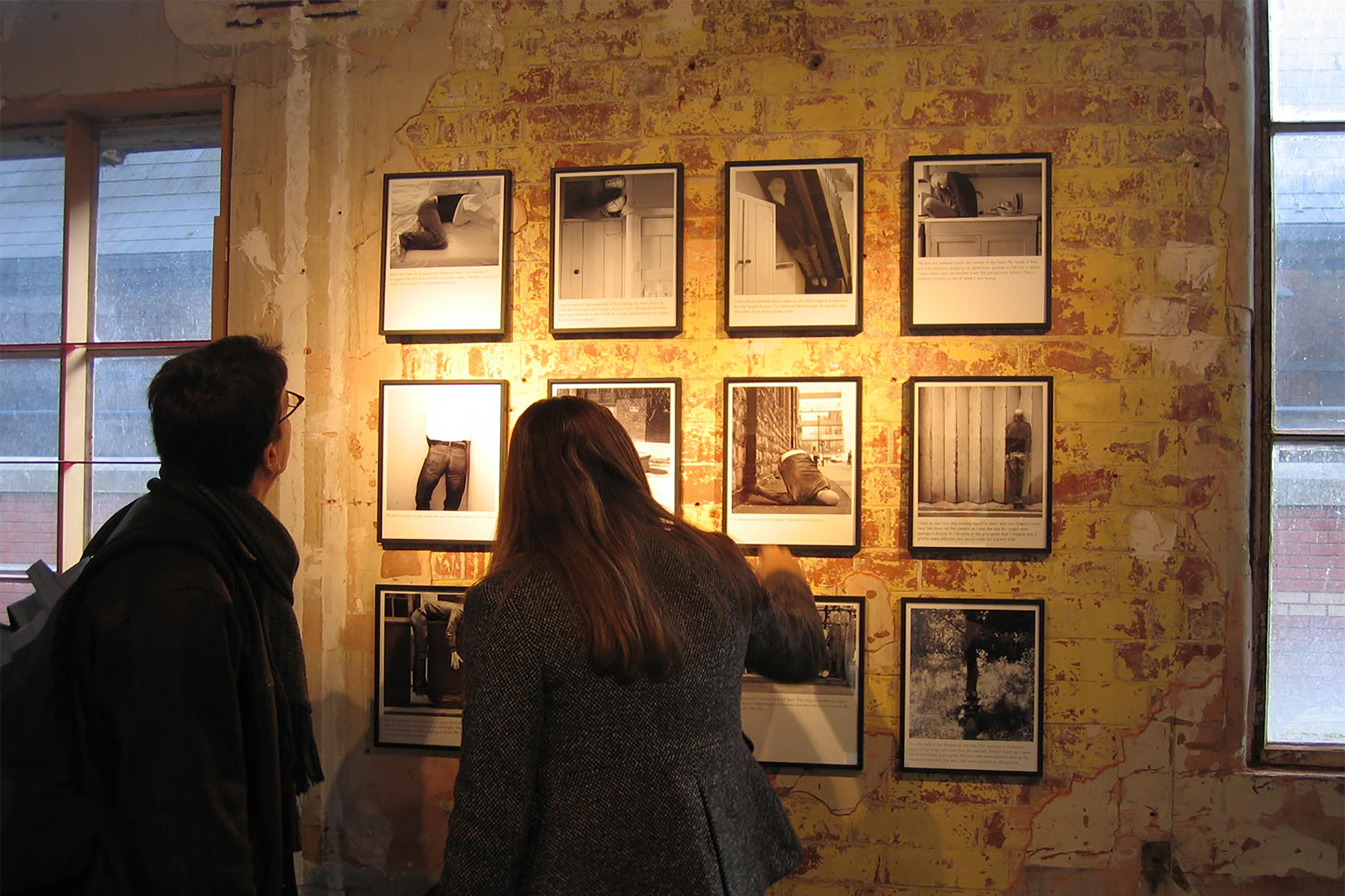 A photograph of a man and a woman observing a wall full of black & white photographs in black frames.