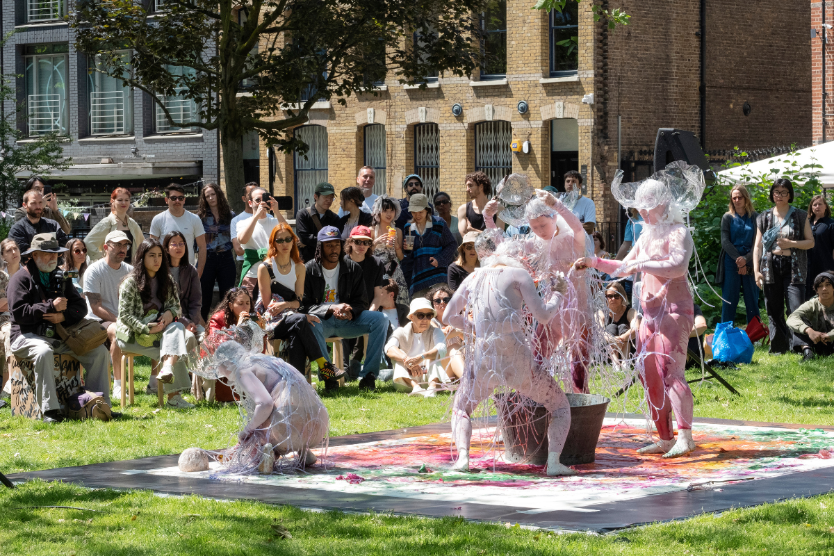 Four performers in white mesh body suits covered in plastic tubes and plastic hats, standing in a sqaure in a dimly lit outdoor square with audience watching them.