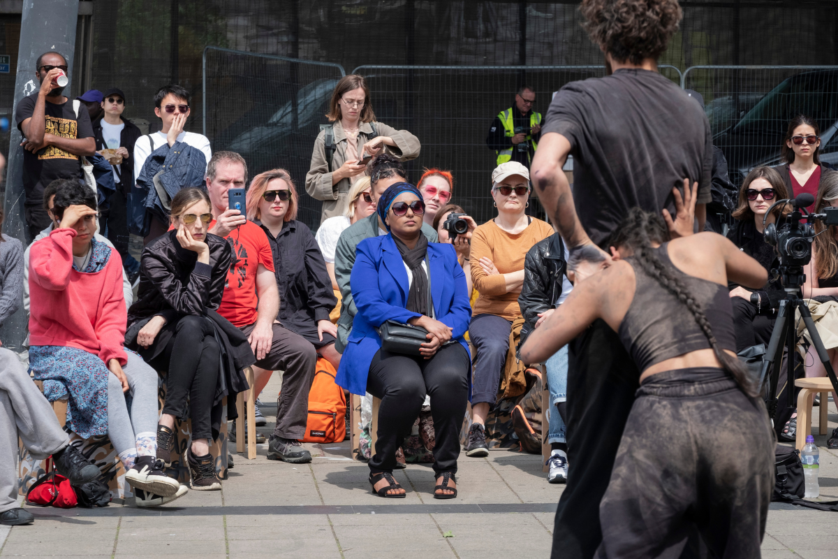 A group of people watching a performance of dancers in black clothing covered in sand in a street.