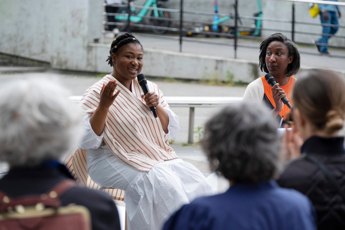 A woman in a white long dress holding a microphone. She is speaking to a group of people.