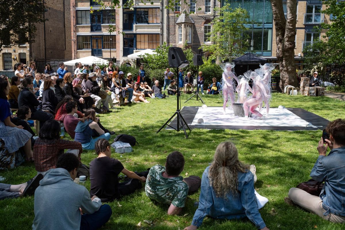 Four performers in white mesh body suits covered in plastic tubes and plastic hats, pouring colourful liquid around from a silver tub. The performers are standing on a large flooring sheet in a park surrounded by a large audience.