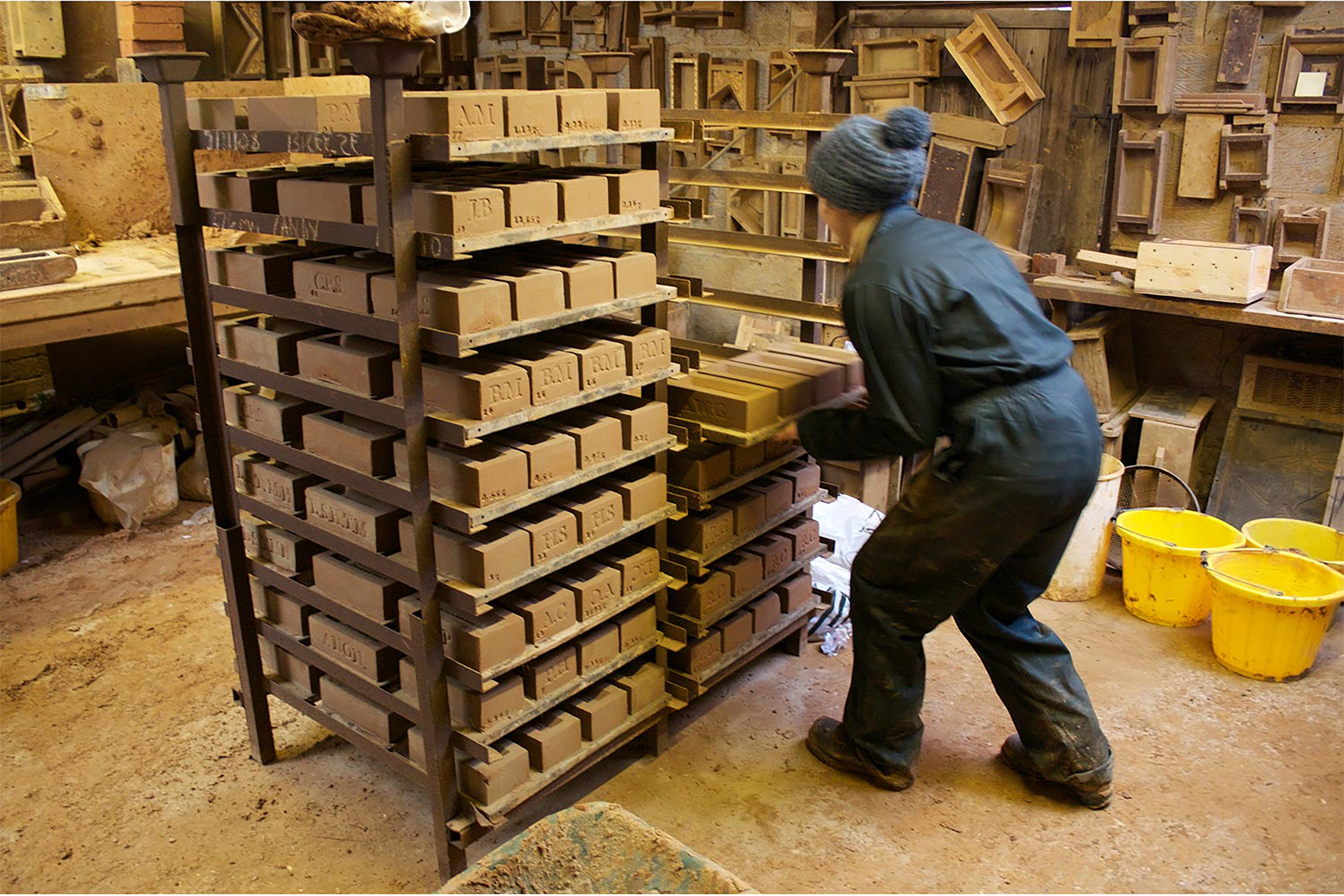 A photograph of a woman wearing a blue jumpsuit puts bricks on a rack inside a studio.
