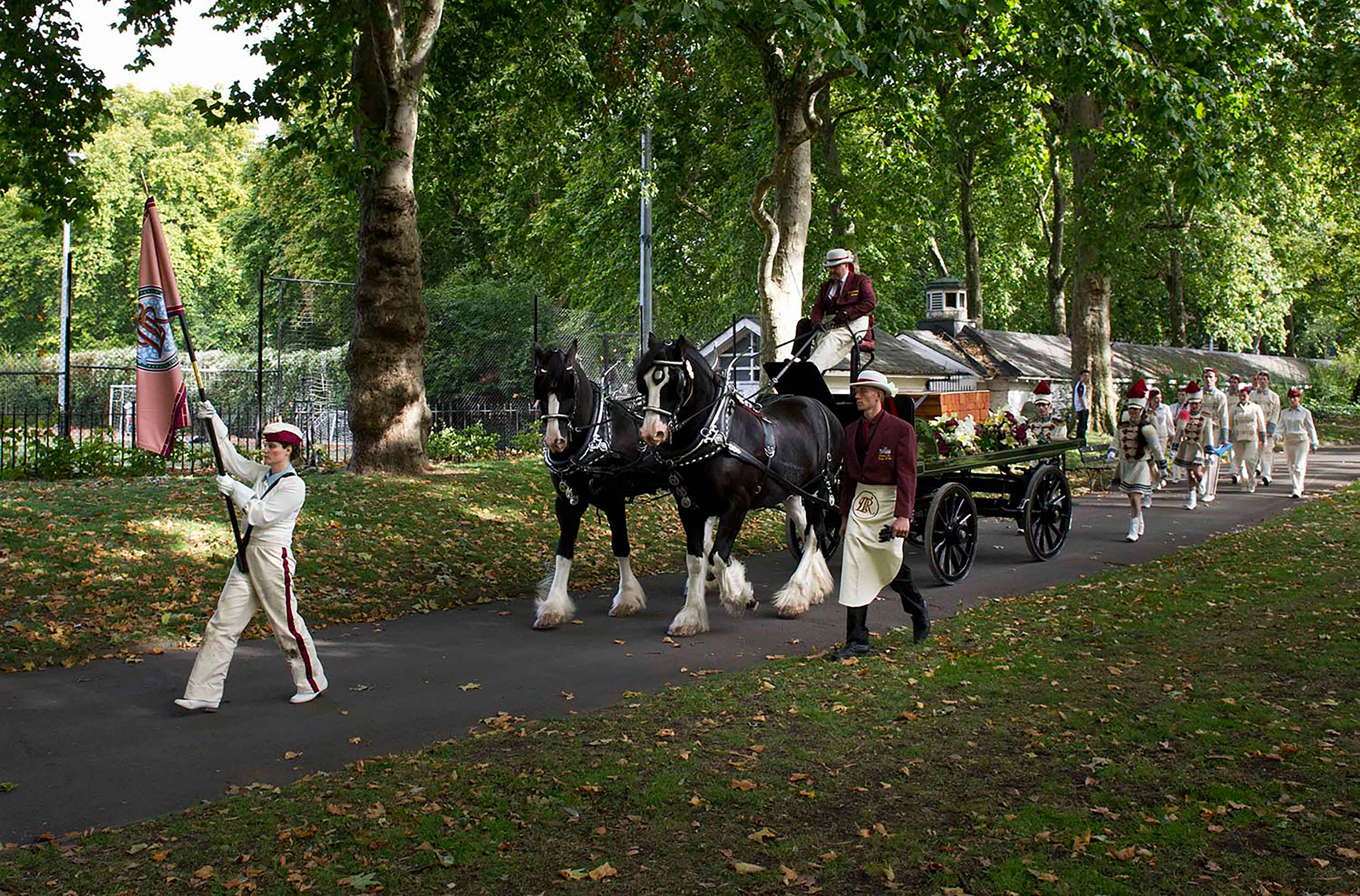 A photograph of a woman holding a banner with a horse and cart behind her marching through a local park. 