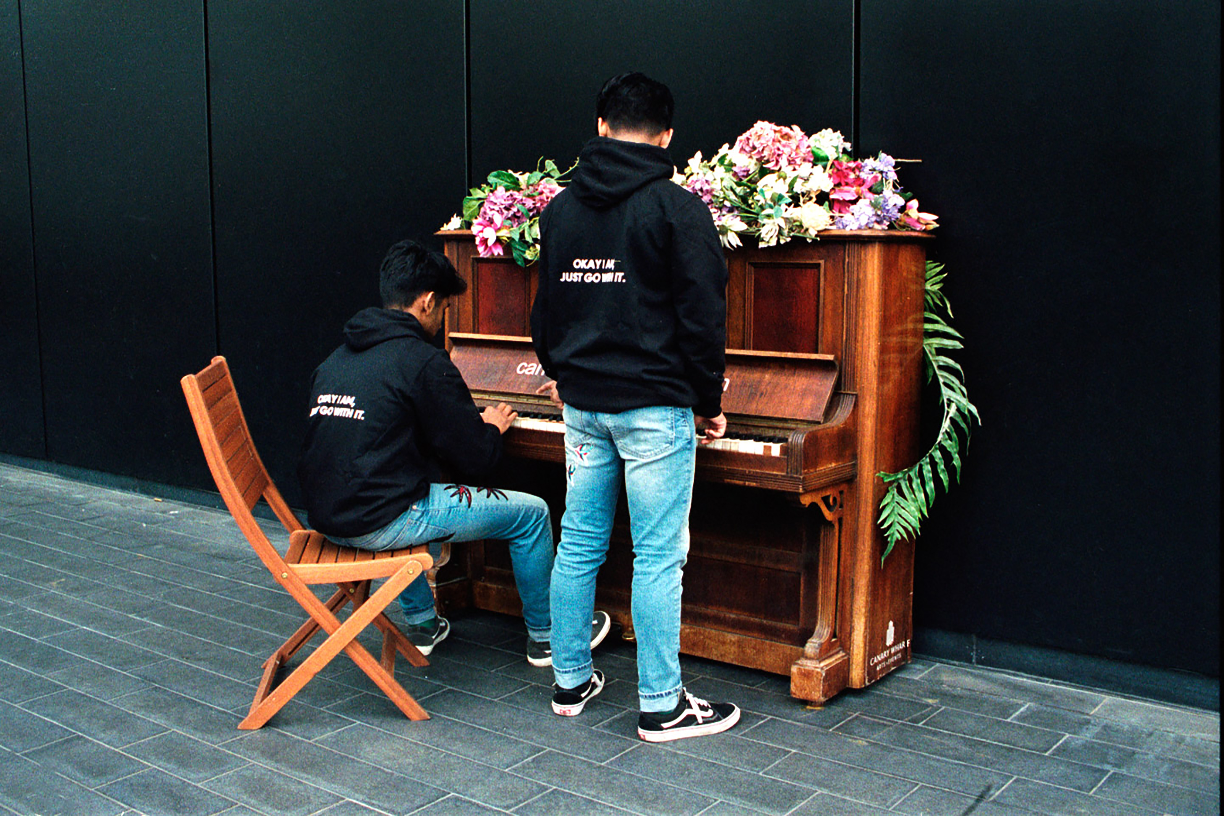 Two young boys wearing black jumpers printed with the words “Okay I am, just go with it” and blue jeans standing in front of an old wooden piano while one plays it.