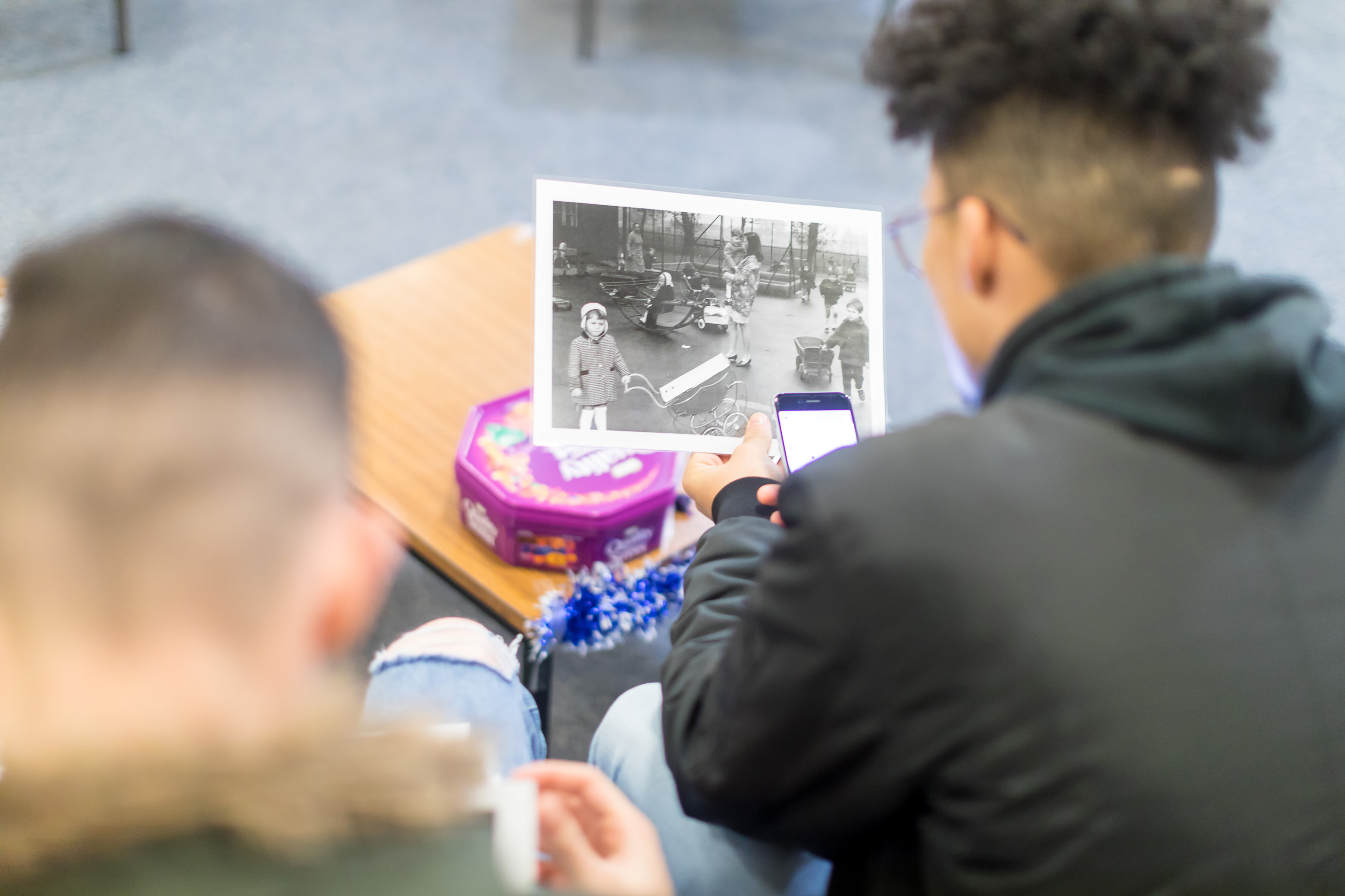 Two young boys from behind sitting on a sofa, with one holding up an old black and white photography of people on a street, in a bright room.