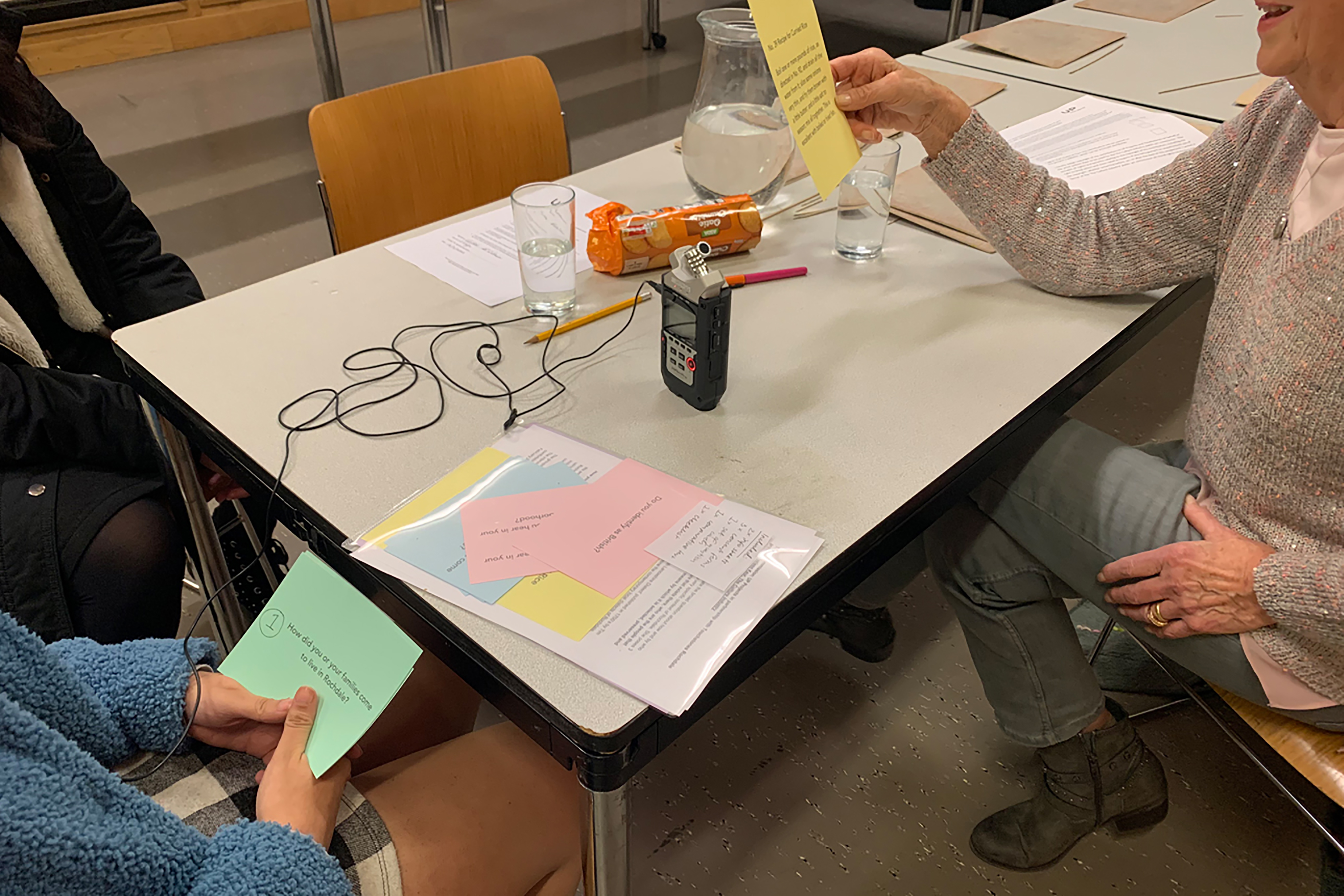 A grey table with a large wire, recording device and colourful pieces of paper, whole two people hold the sheets and ask each other questions.