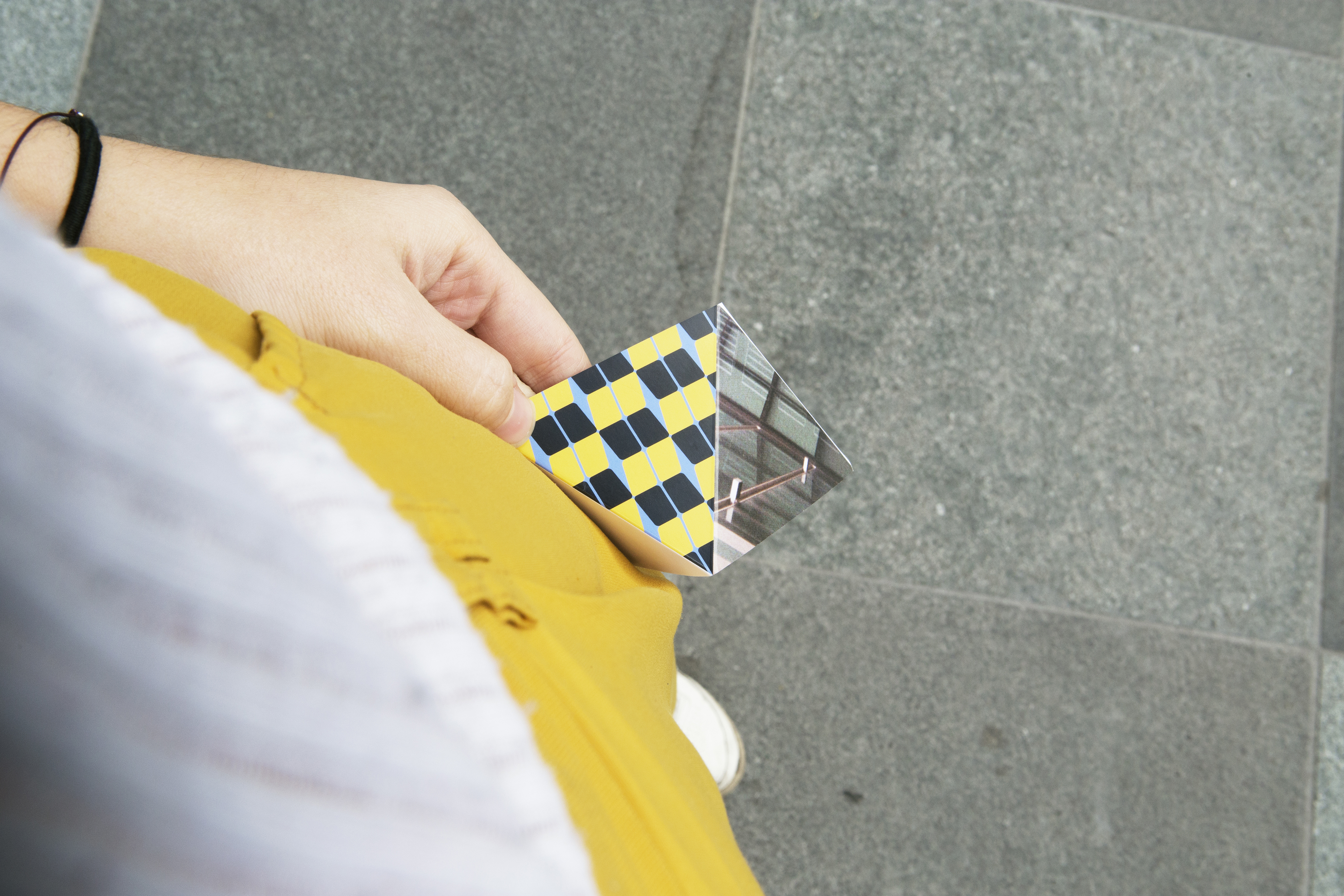 A person holding a folded piece of paper made up of colourful pattered blocks, against their hip standing on the street.