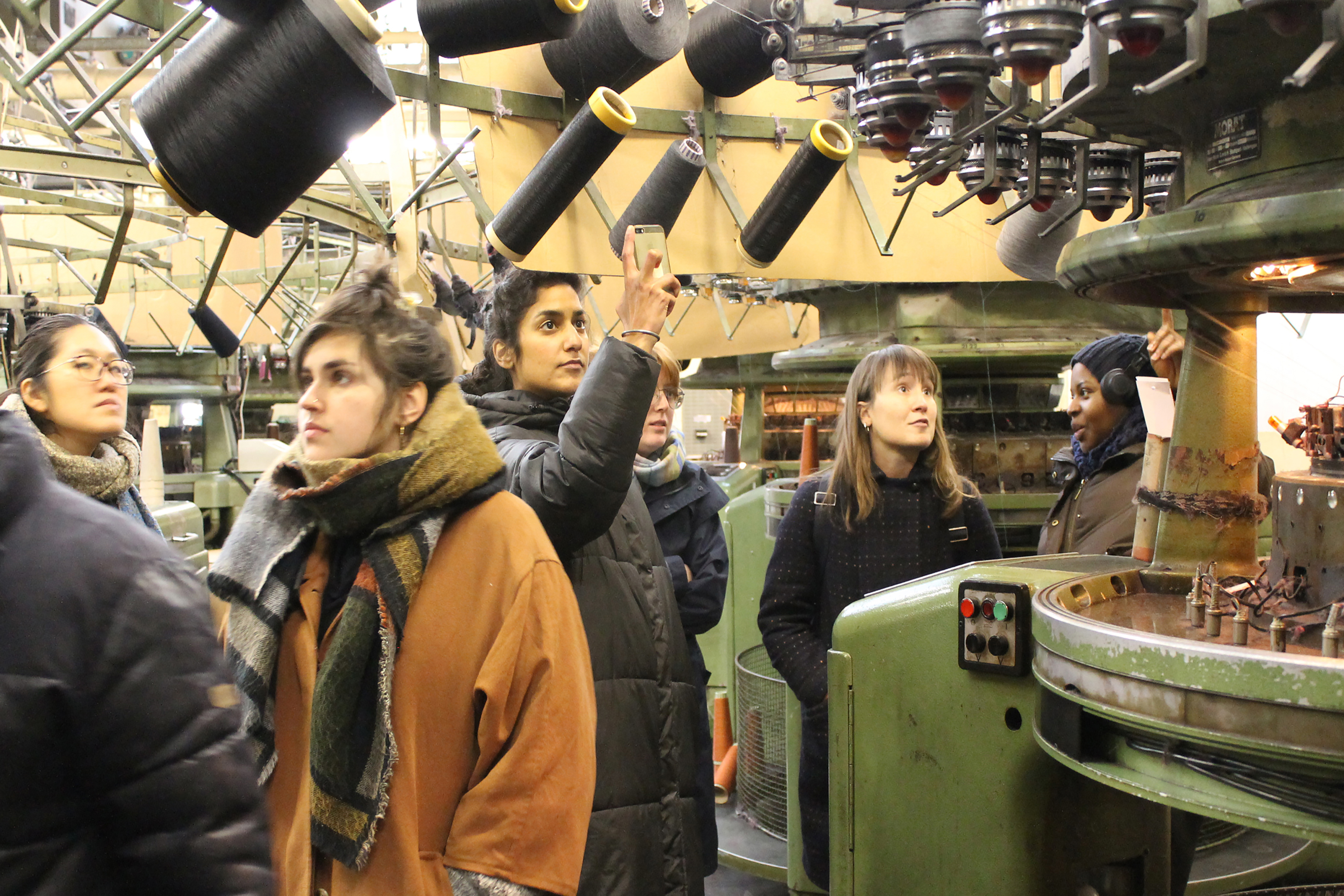 A group of people wearing coats and scarves, standing and observing a fabric factory machinery.
