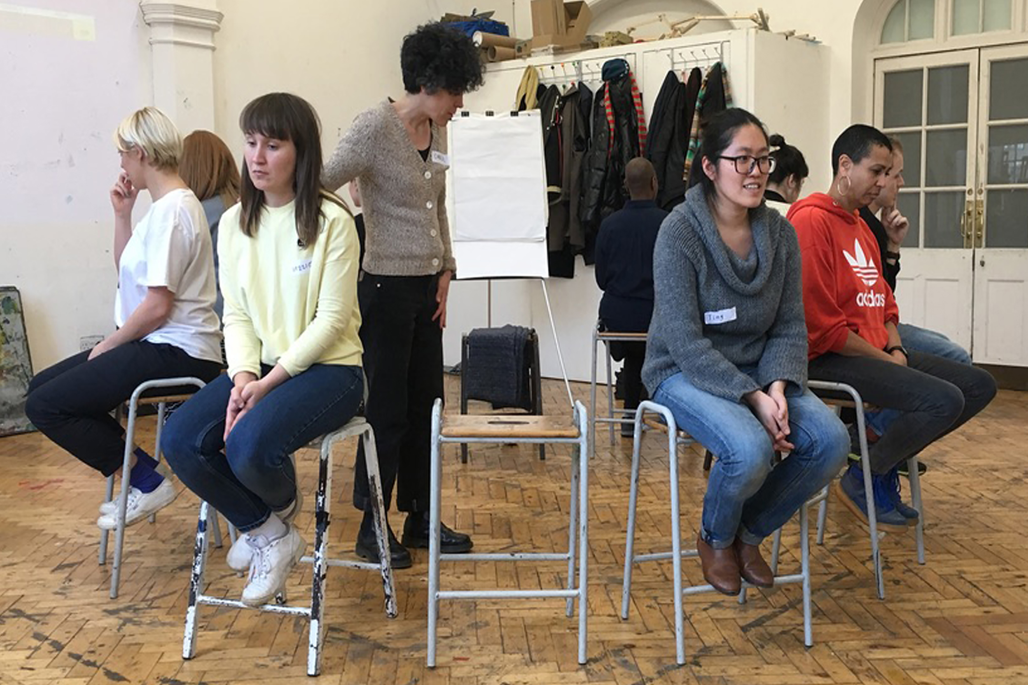 A group of people sitting on stools facing away from each other forming a circle in an open hall with a wooden floor.