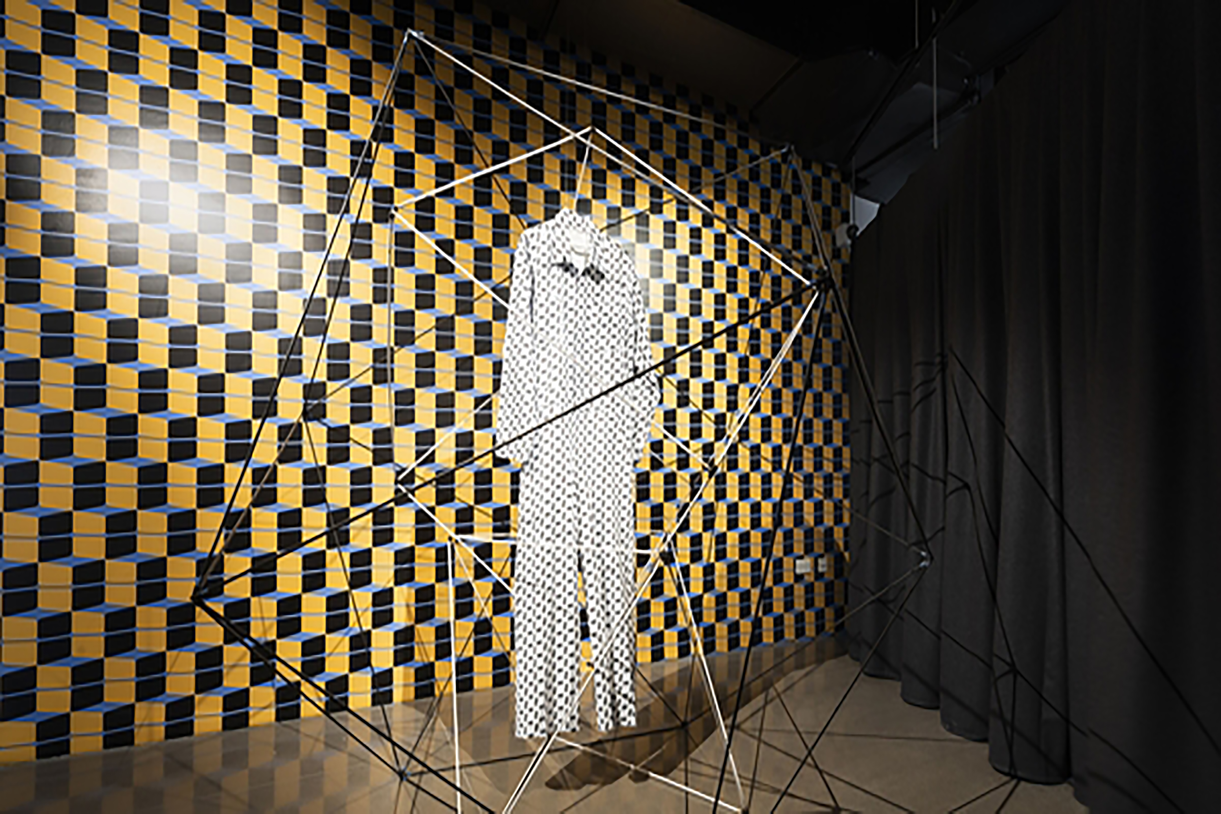 A white and black geometric patterned outfit handing from a metal structure in a gallery in front of a geometric background.