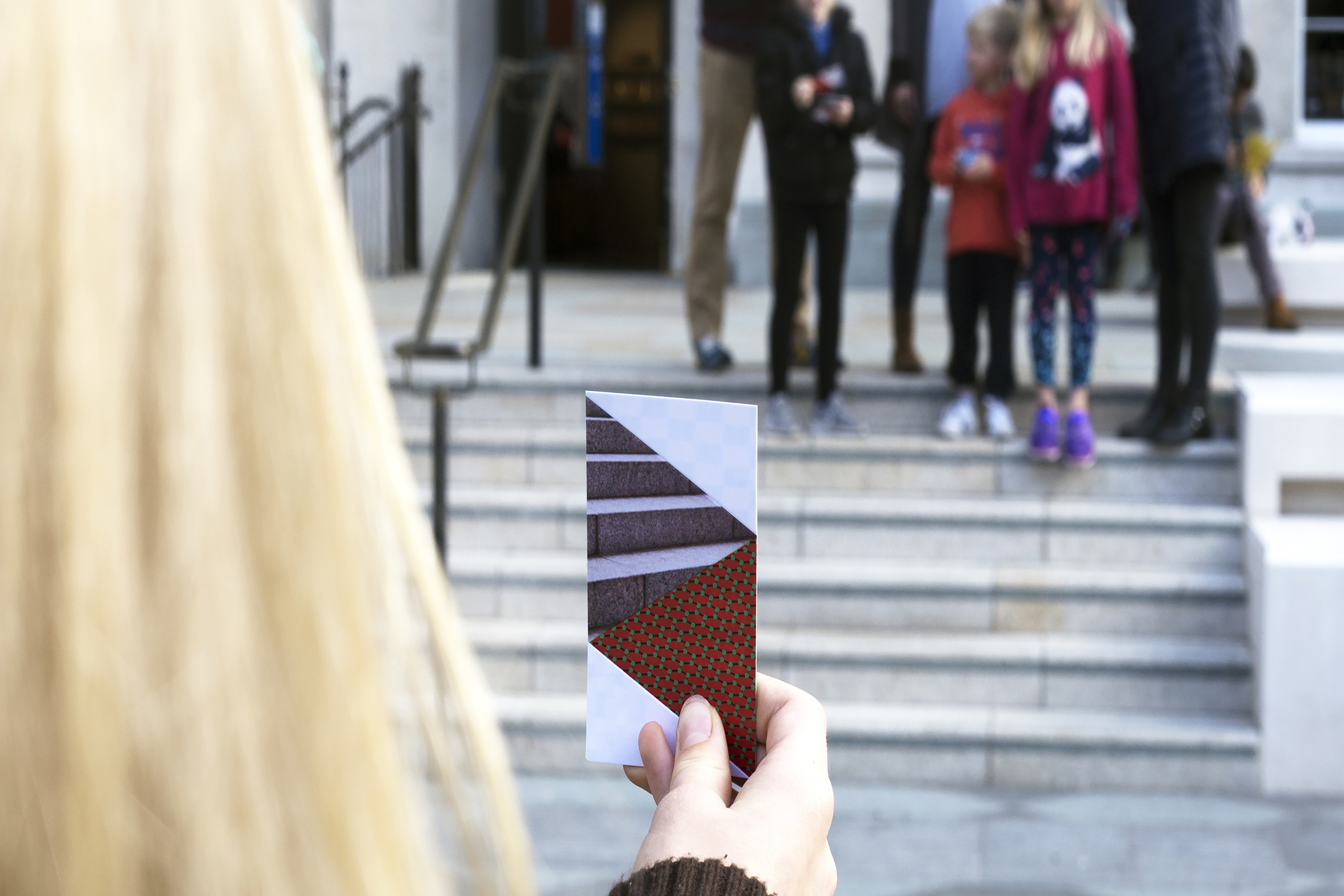 A person holding up a card with a geometric print on it, in front of a staircase outside of a gallery.