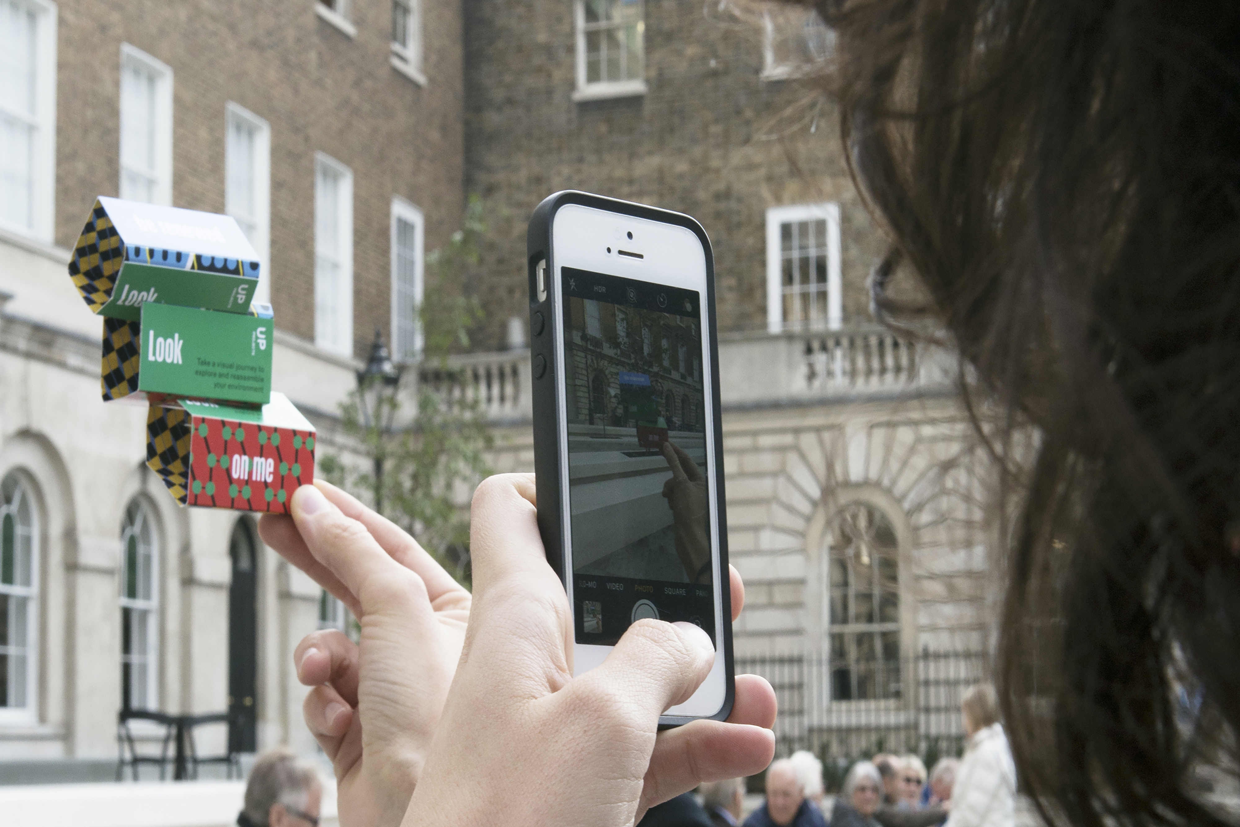 A person holding up a card with a geometric print on it, in front of a staircase outside of a gallery, while taking a picture on their phone.