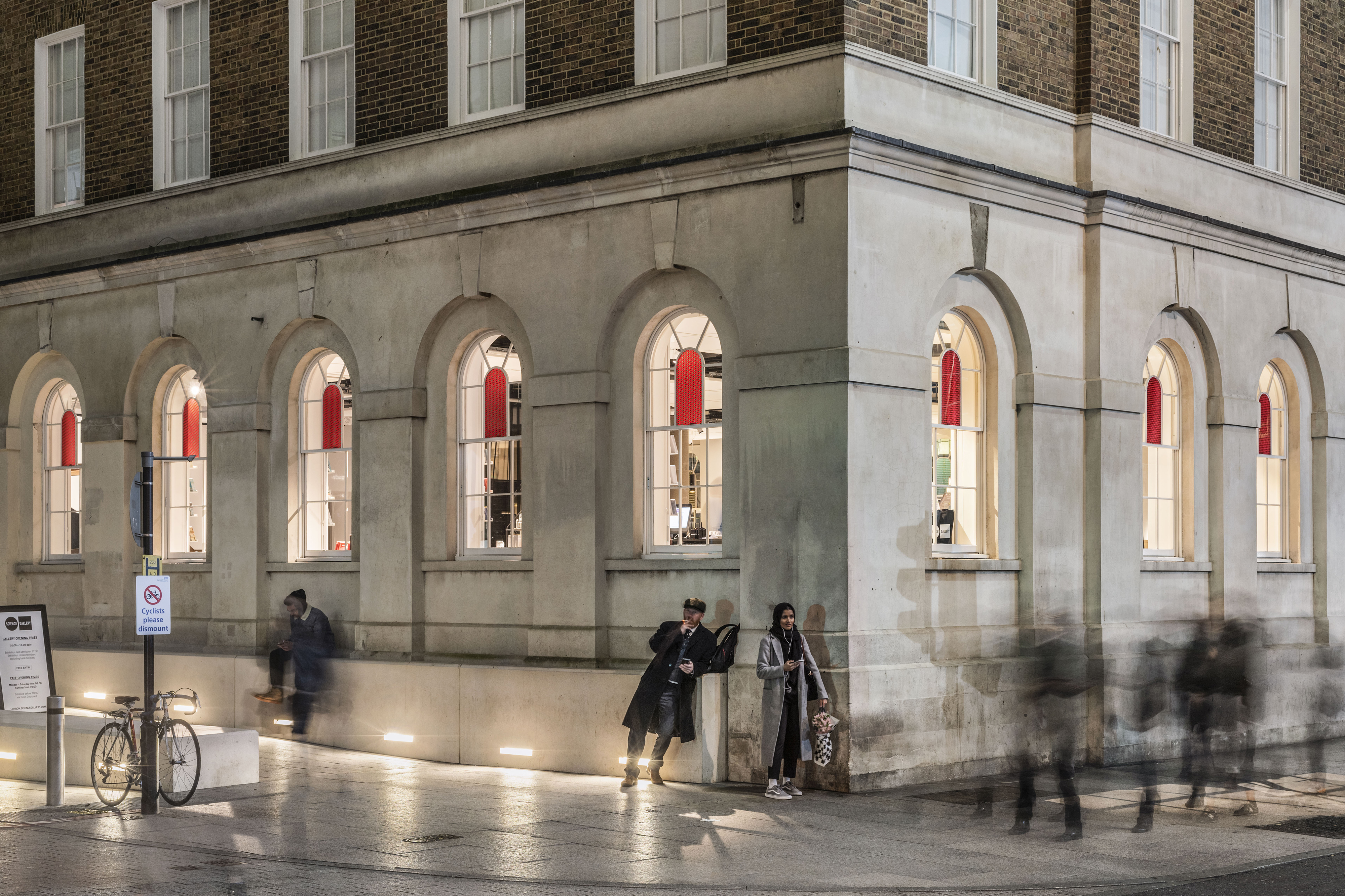 Outside of a gallery with white walls with large, coloured stain glass windows at night.