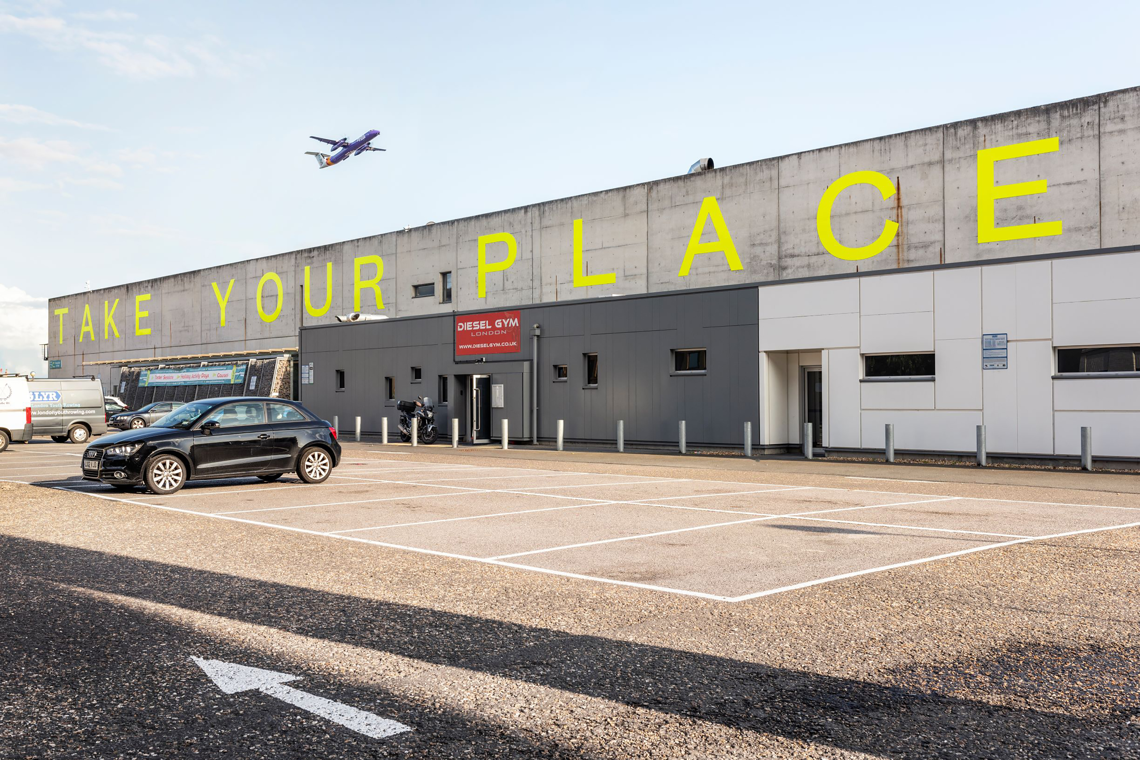 Large neon yellow letters that spell out the words "Take Your Place" situated at the top of a large concrete grey building, next to a car park, with a plane flying over.