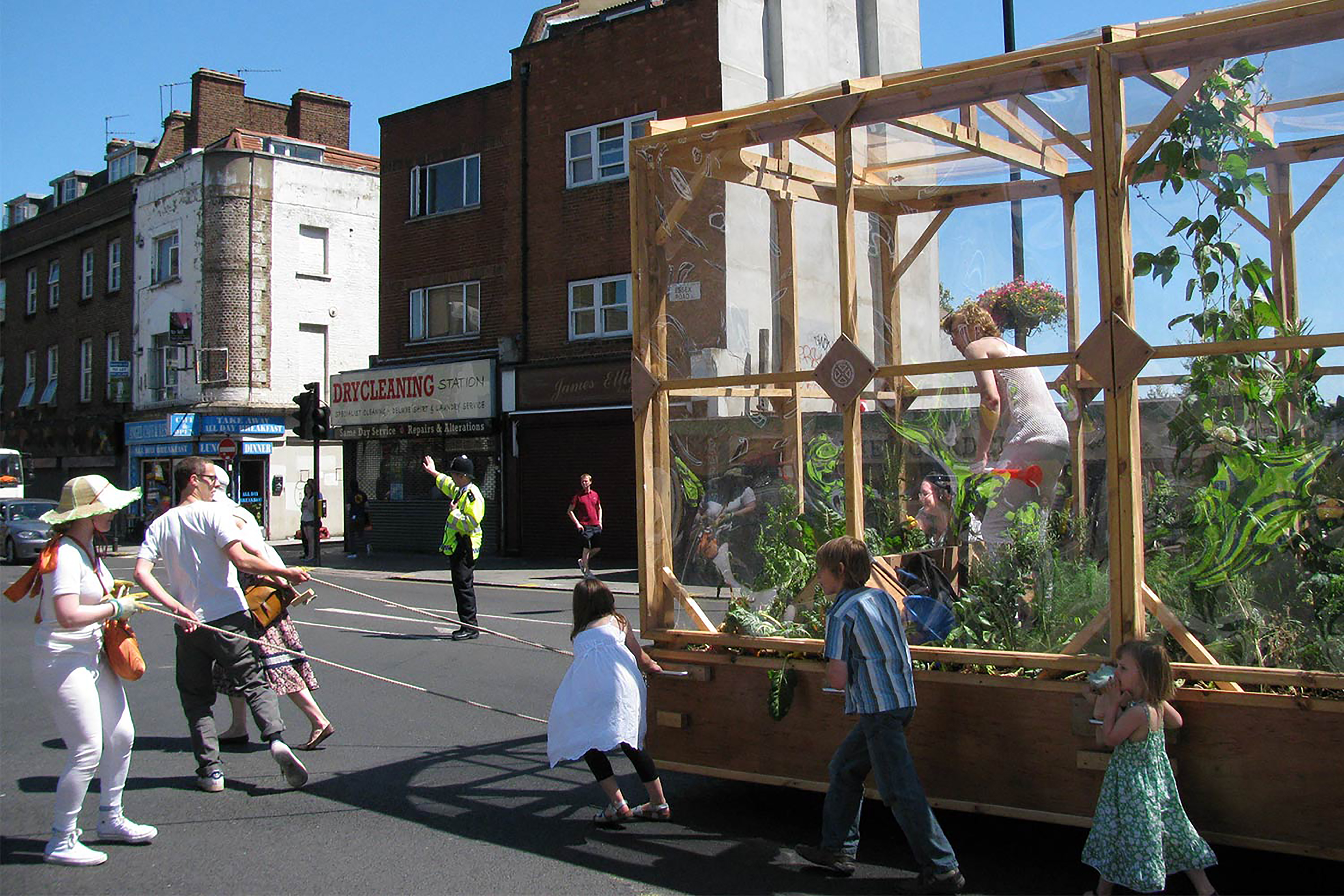 A photograph of a family pulling along a wooden framed green house with a woman inside in the middle of the road.