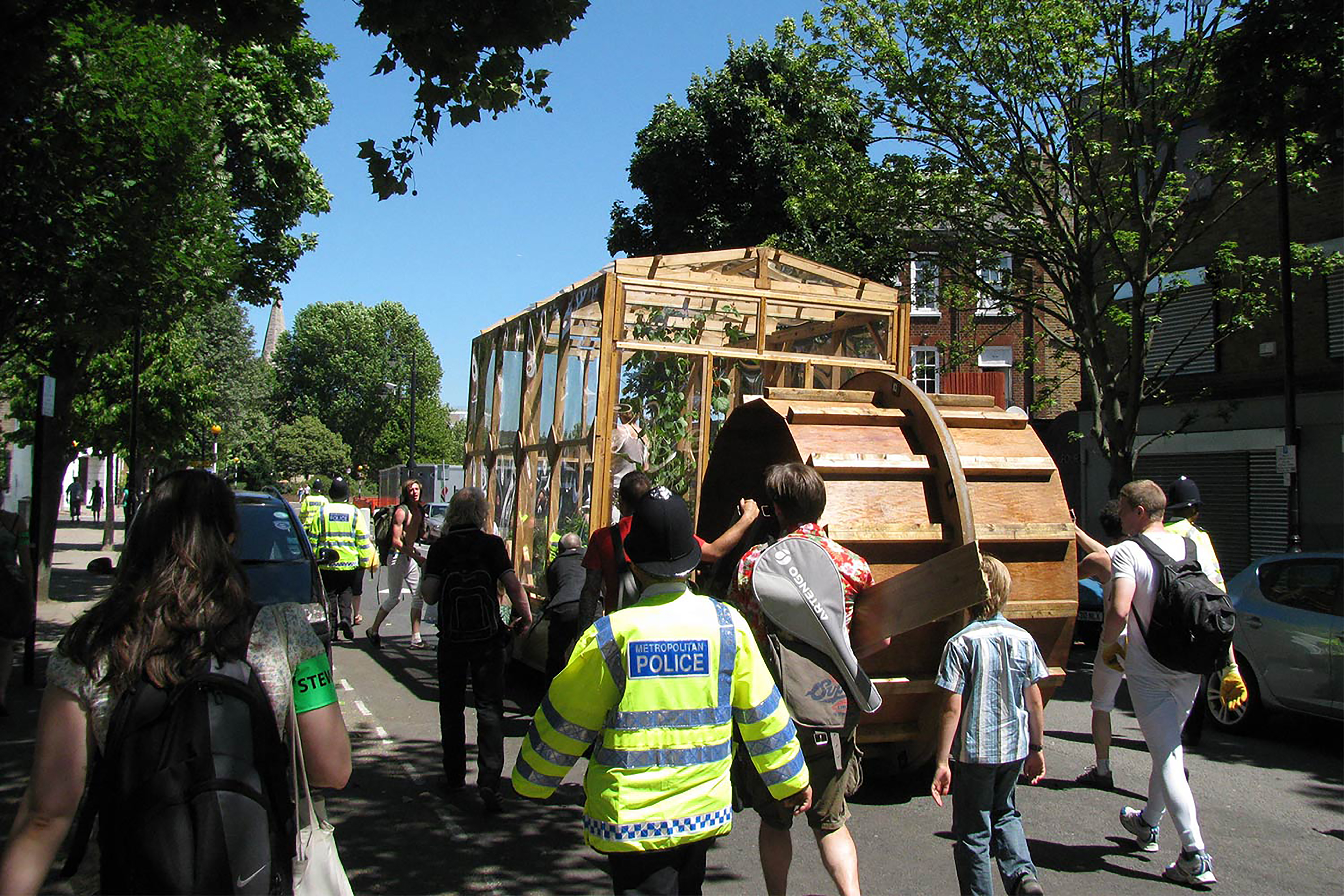 A photograph of a parade of people pulling along a wooden framed green house in the middle of the road.